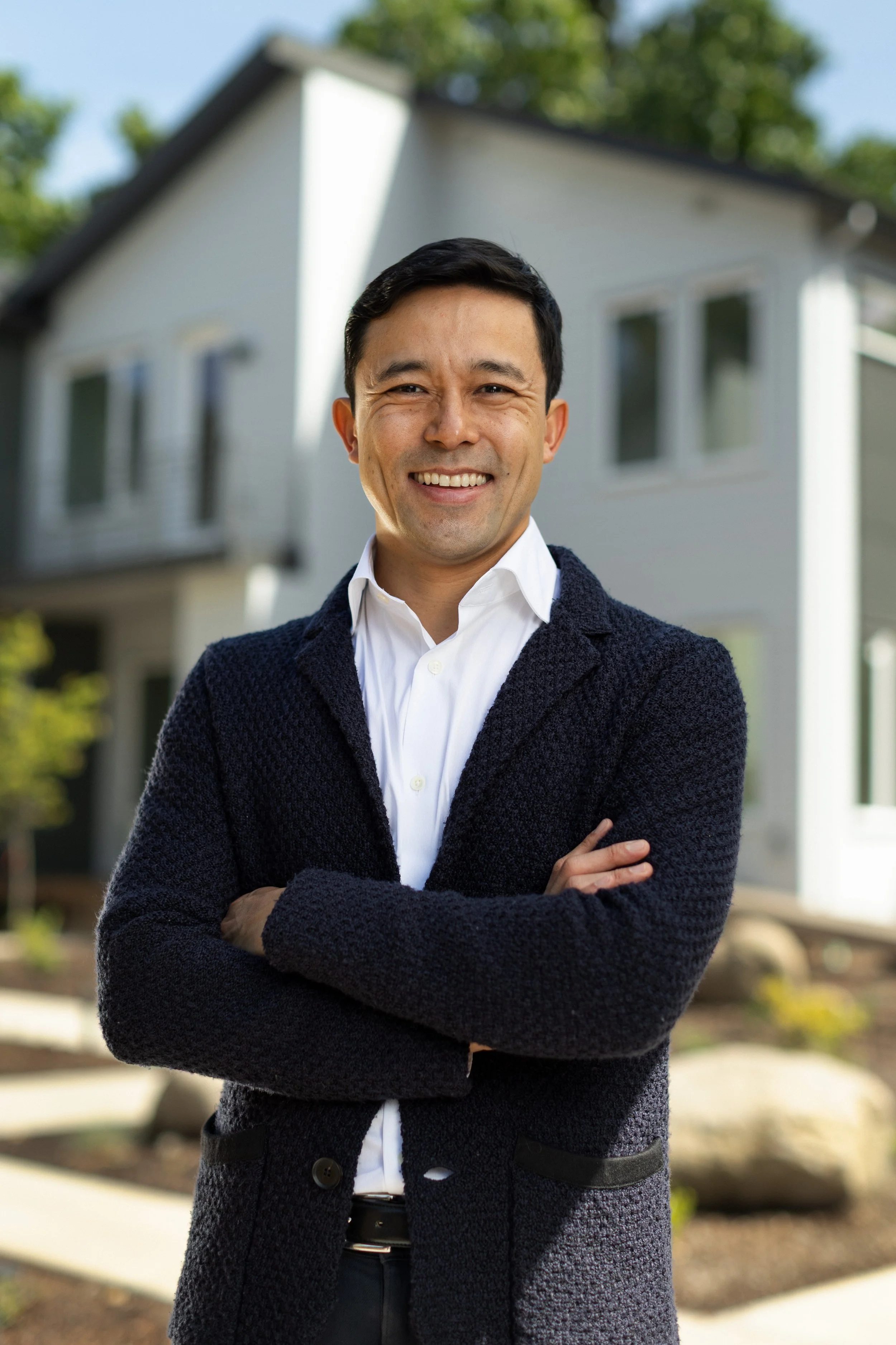 Man smiling, standing outdoors in front of a house with trees, wearing a white shirt and a dark blue cardigan, with arms crossed.