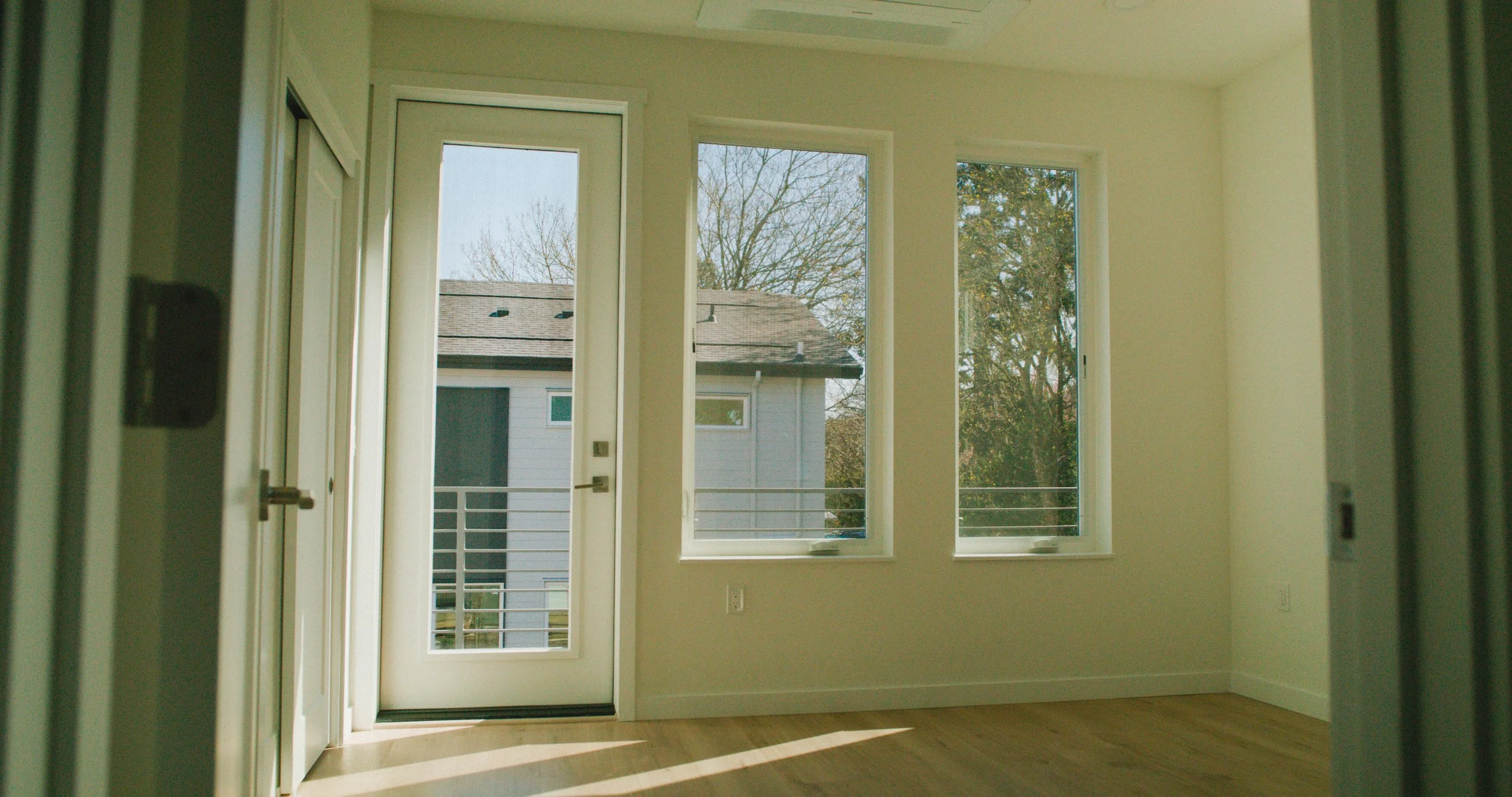 Empty room with large windows and a door leading to a balcony, showing a view of neighboring houses and trees outside.