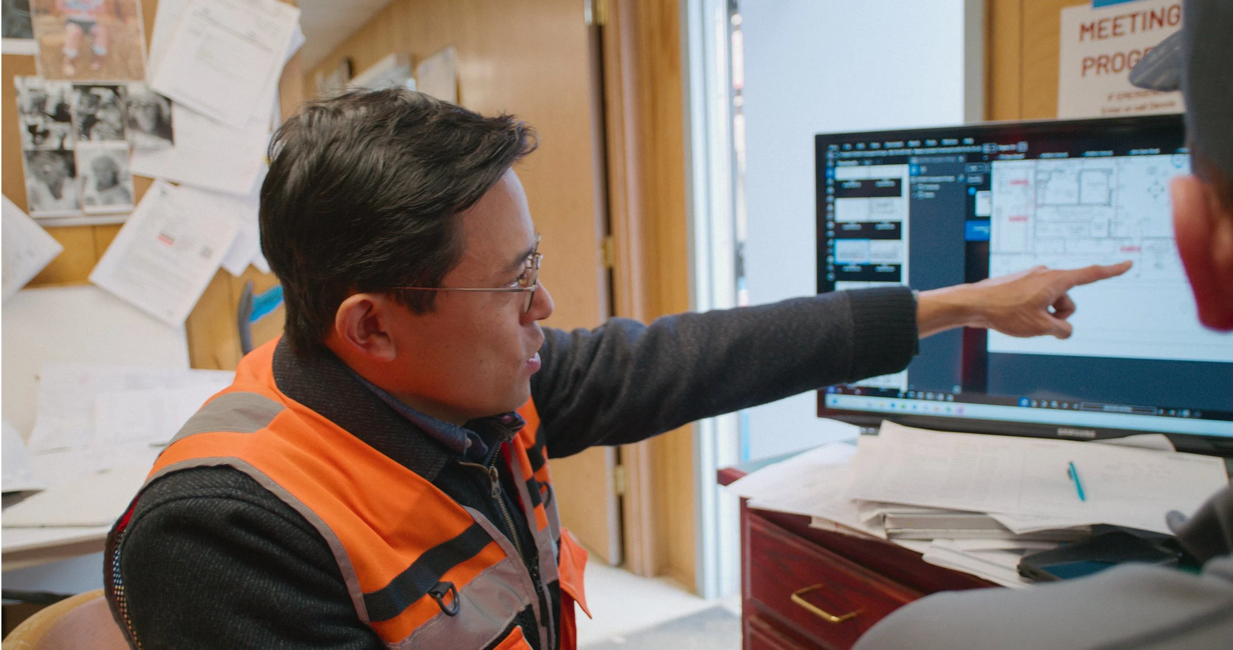 A man wearing glasses and an orange safety vest pointing at a computer monitor in an office or control room.