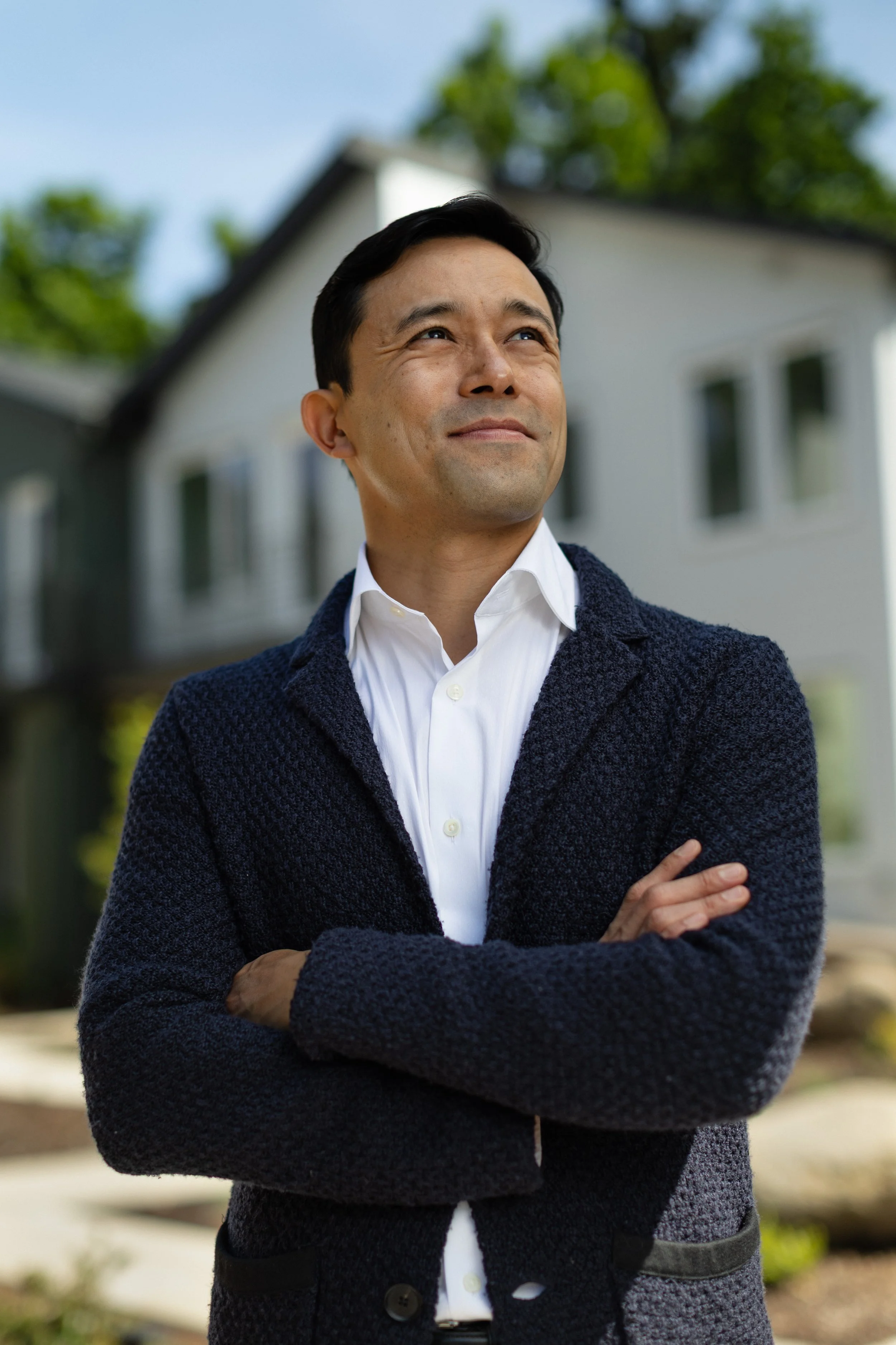 A man with short black hair and light skin, wearing a white shirt and dark blue textured sweater, stands outdoors with arms crossed, smiling and looking slightly upward. There is a house with white walls and windows behind him, surrounded by greenery and trees, under a clear sky.