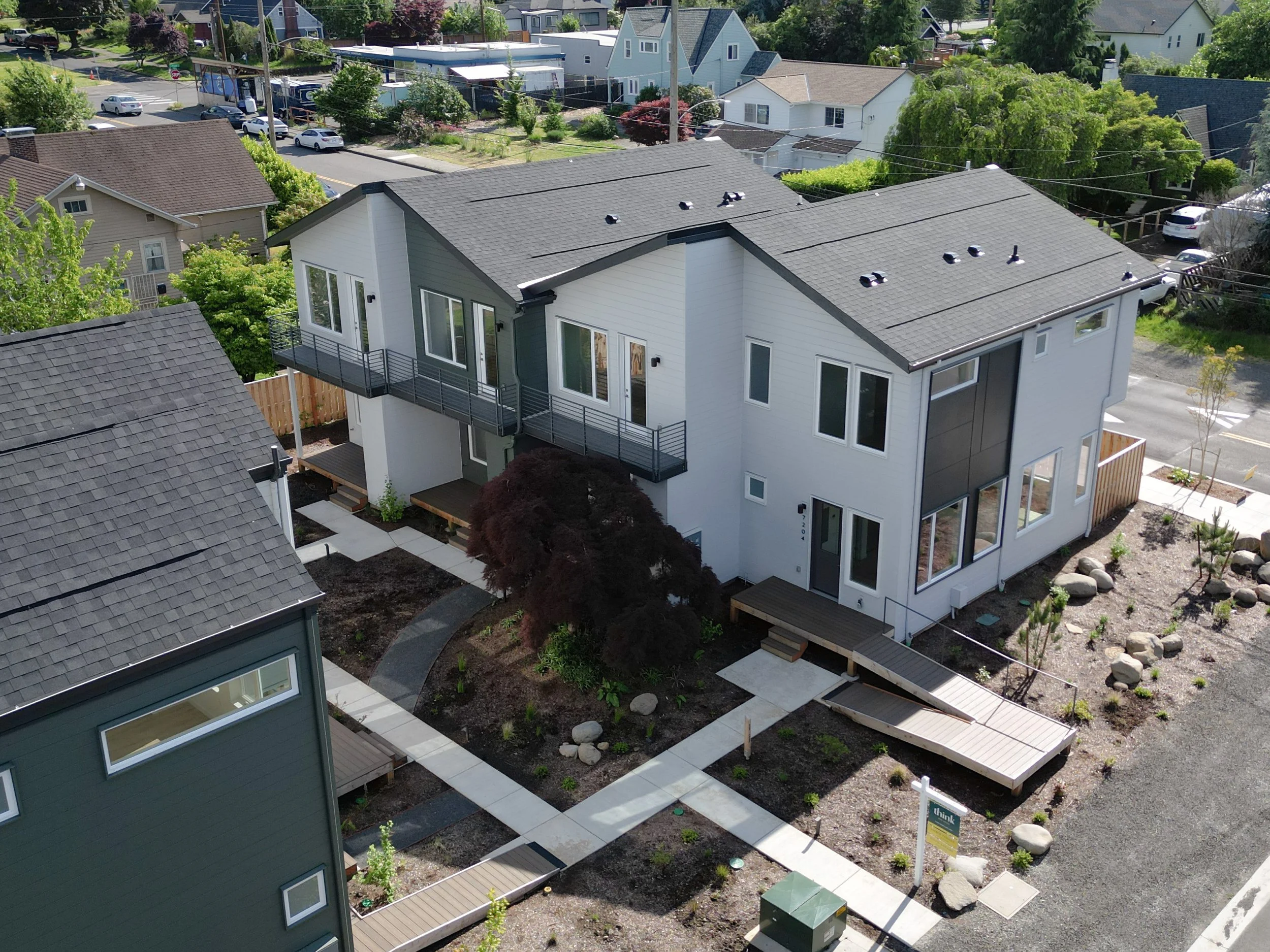 Aerial view of a modern two-story house with a gray roof and white exterior, surrounded by a landscaped yard with young trees, shrubs, benches, and paved walkways, in a residential neighborhood with other houses and parked cars.