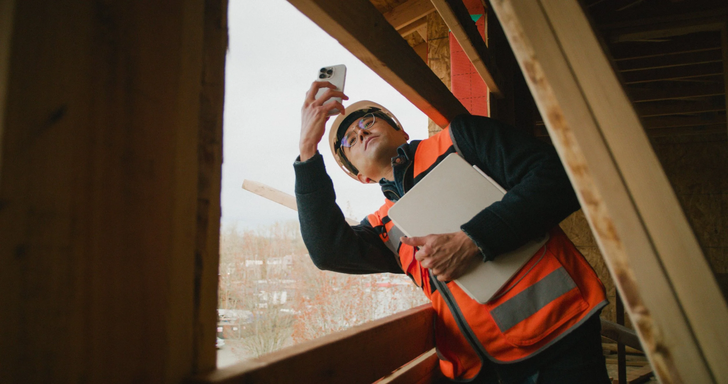 A construction worker wearing safety glasses and a helmet, holding a tablet and smartphone, is looking up inside a wooden construction frame.