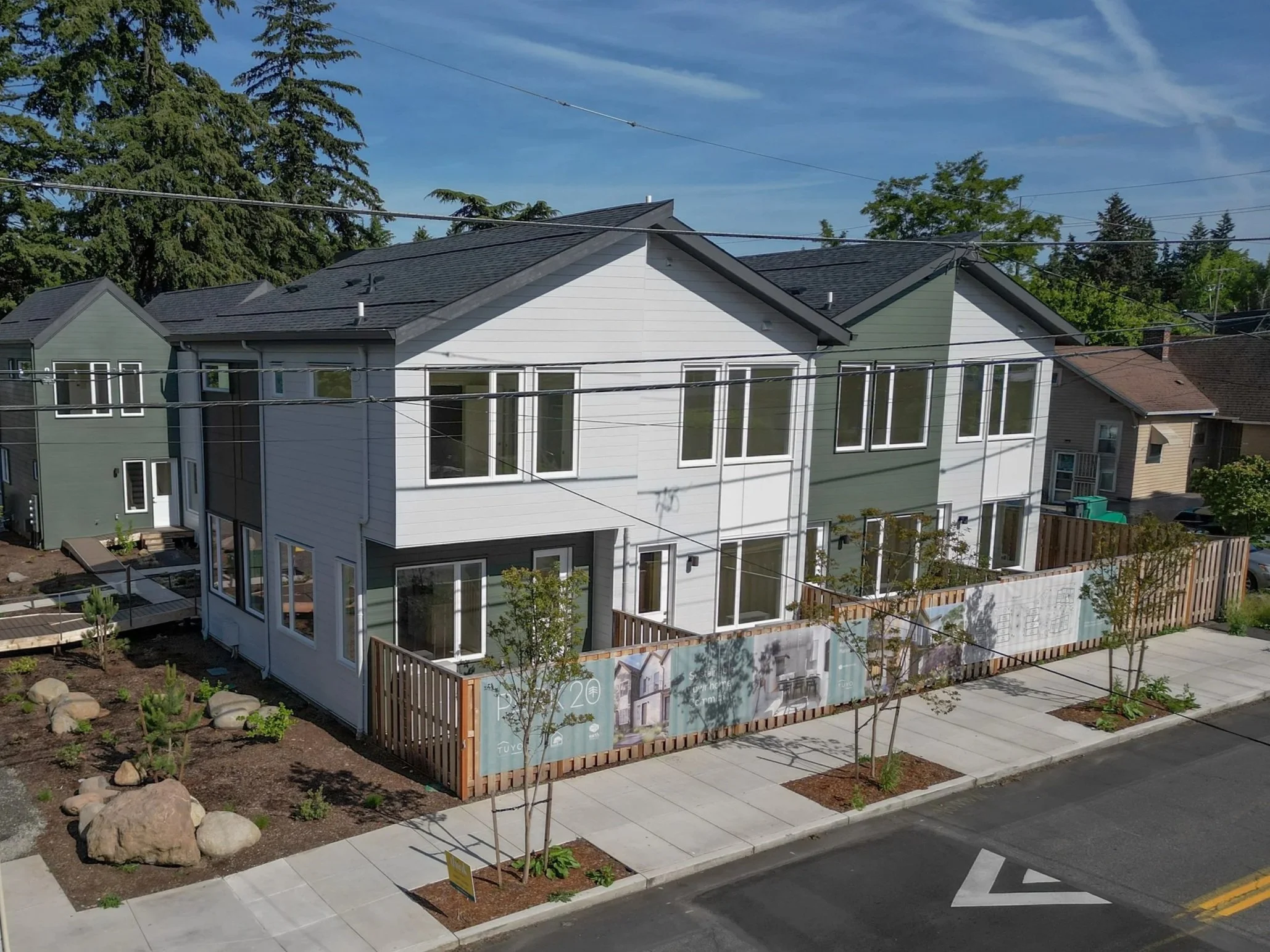 Newly constructed multi-story residential building with large windows, a gray and white exterior, and a small backyard. The building is behind a sidewalk with young trees and some rocks, and a wooden fence with signage advertising the property.