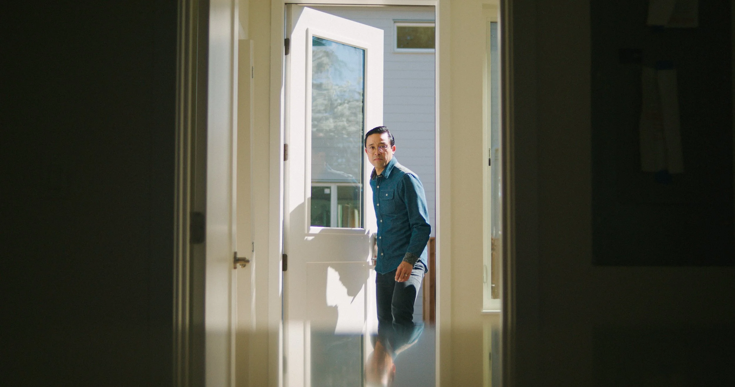 A man in a denim shirt and glasses standing at the open back door of a house, looking inside.