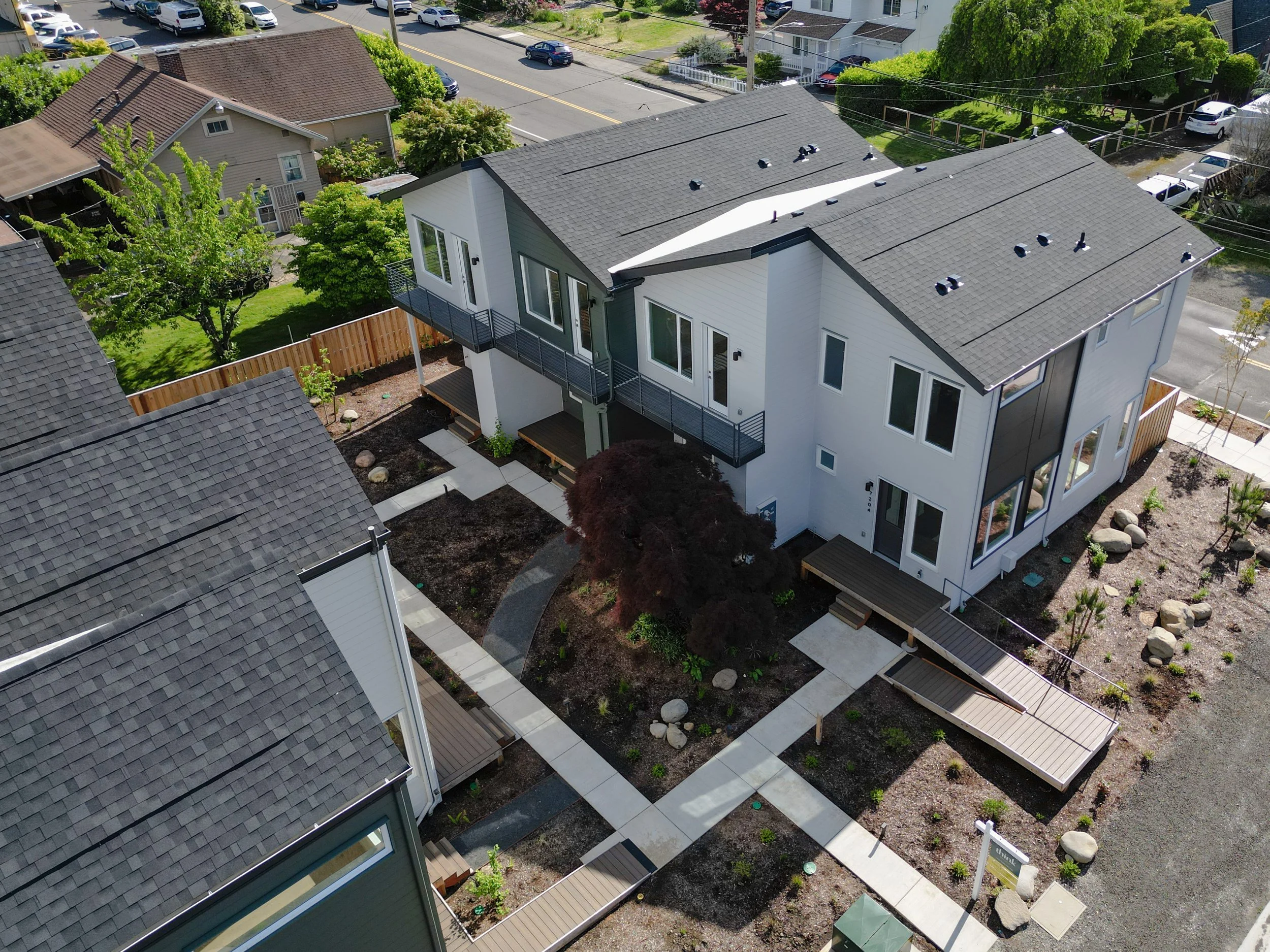 Aerial view of a modern residential building with a gray roof, white and gray exterior walls, large windows, a balcony, and landscaped backyard with a path, trees, and decorative rocks.