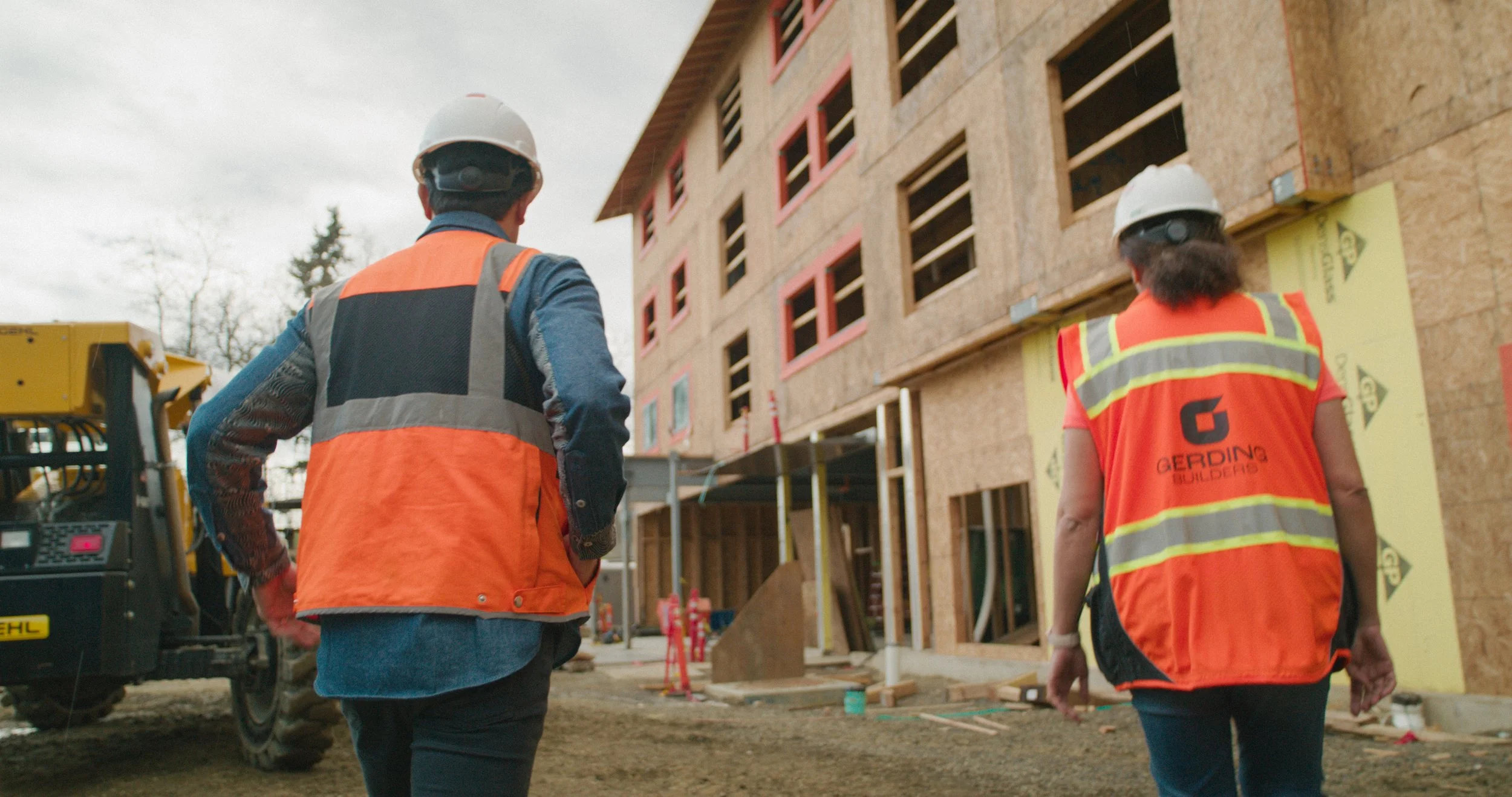 Two construction workers, one man and one woman, walking towards a multi-story building under construction. They are wearing white hard hats and bright orange safety vests. The building's exterior is partially covered with yellow sheathing, and there are open window frames. Construction equipment and materials are visible around them.