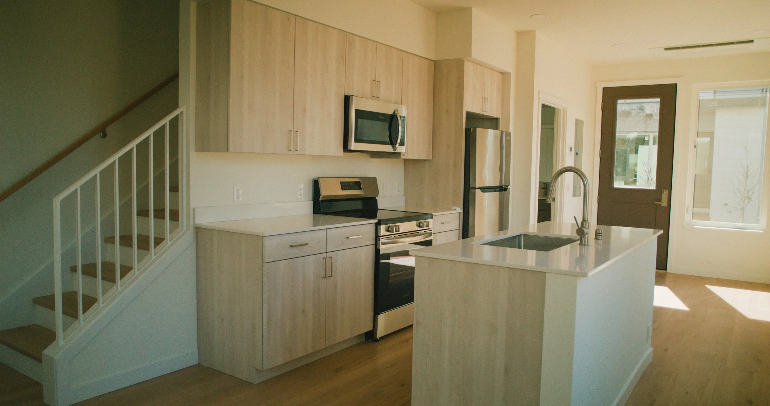 Modern kitchen with light wooden cabinets, white countertops, stainless steel appliances, a central island with a sink, and large windows and door letting in natural light.
