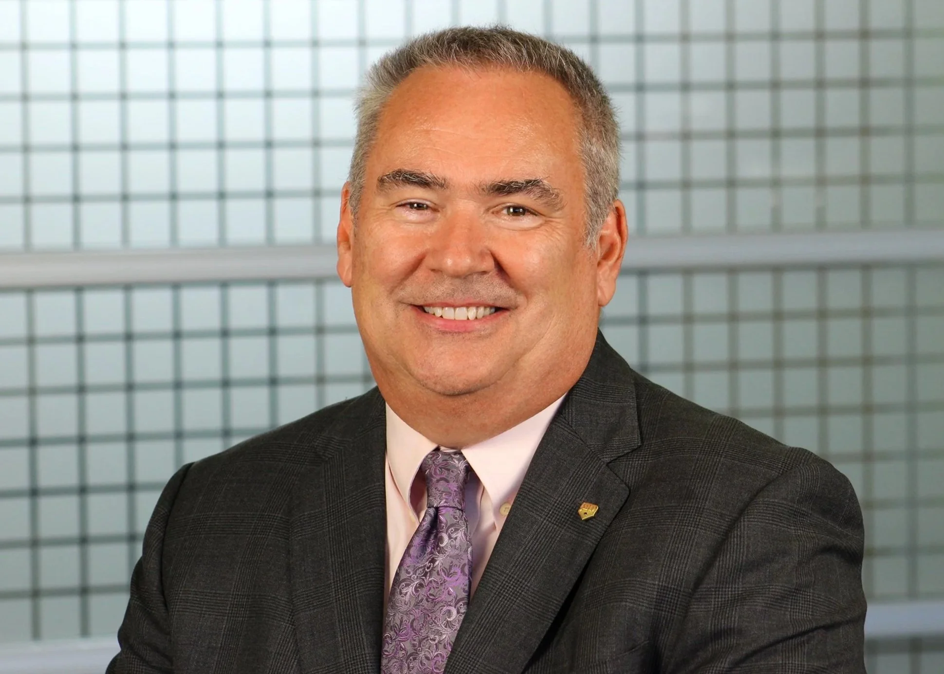 A middle-aged man with gray hair smiling in a professional suit and tie, standing in front of a modern glass building.