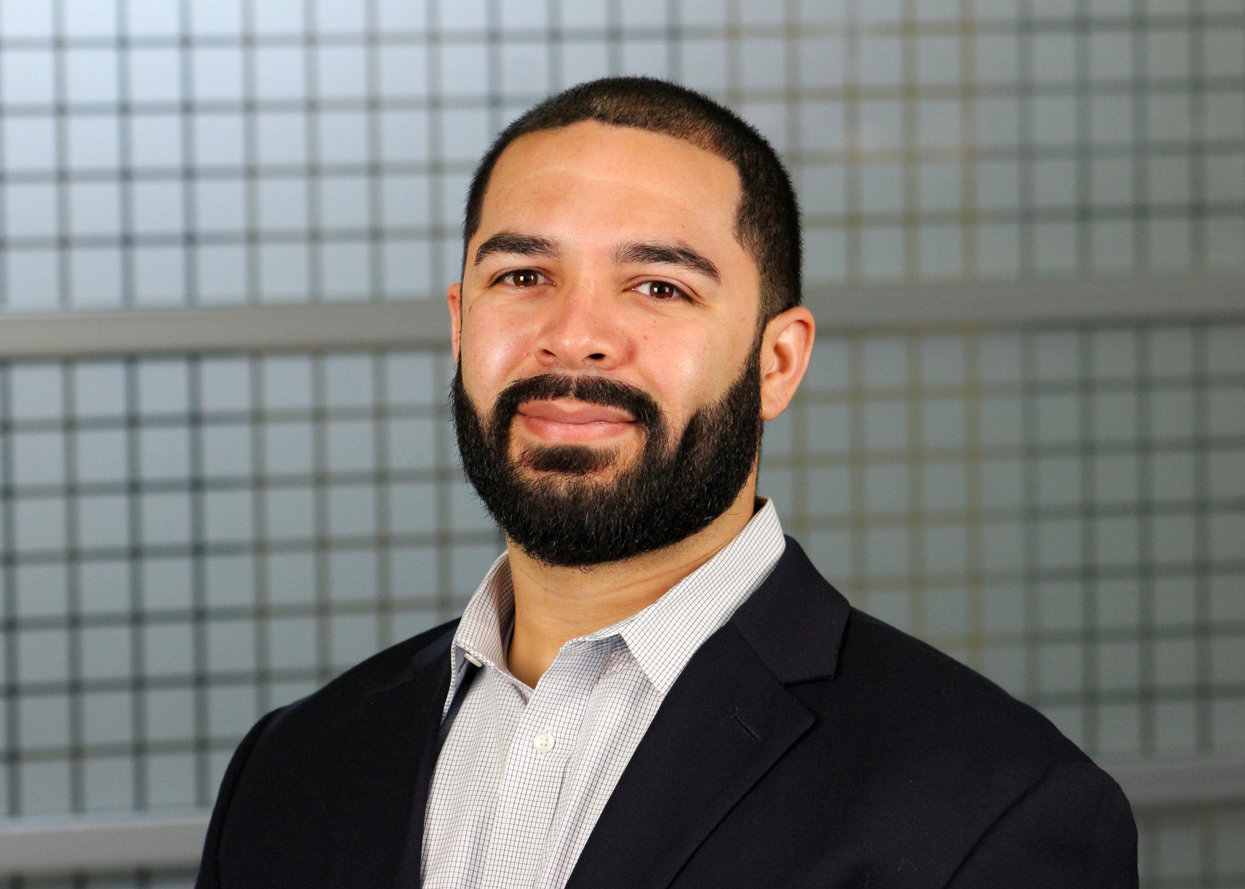 Professional headshot of a man in a business suit, wearing a blue shirt and a gray patterned tie, standing in an office corridor with yellow walls and windows.