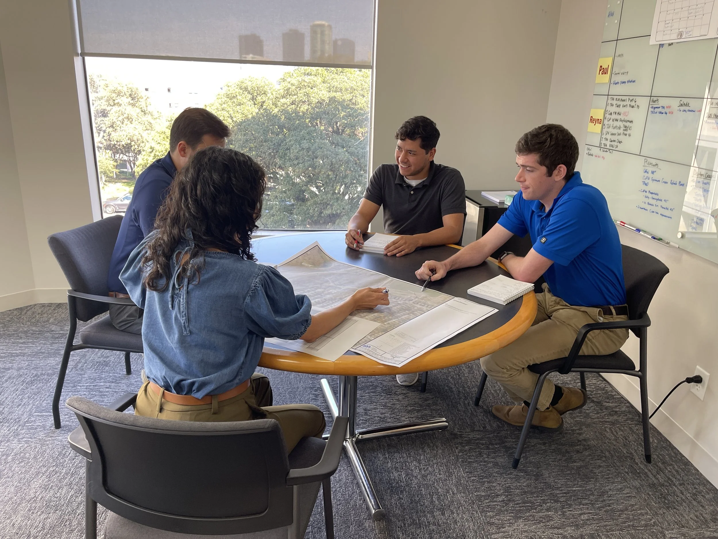 Four young adults in a meeting room sitting around a table with papers, notebooks, and a map, engaged in discussion. A whiteboard with notes and a schedule is visible on the wall. Large windows with a view of trees and buildings outside.