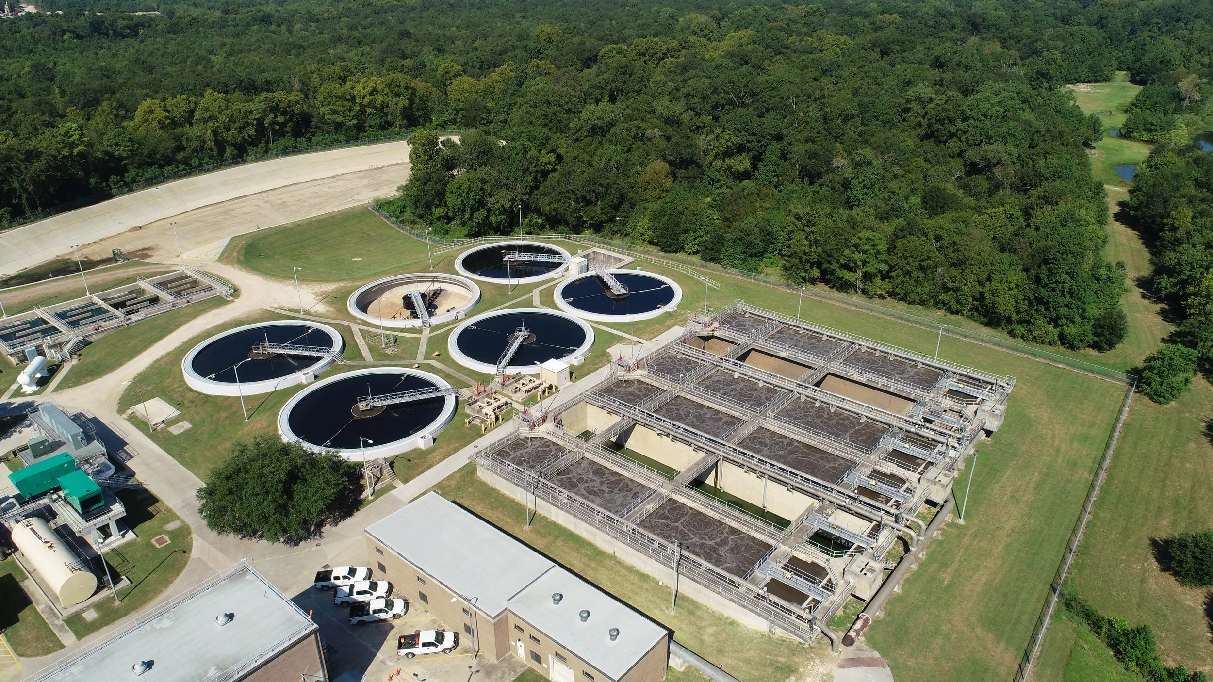Aerial view of a wastewater treatment plant with circular and rectangular tanks surrounded by green trees and grass.