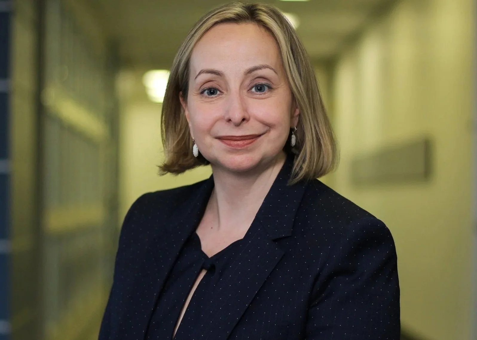 A woman with shoulder-length blonde hair, wearing a black blazer with subtle dots, standing in a brightly lit corridor with yellow and blue walls.