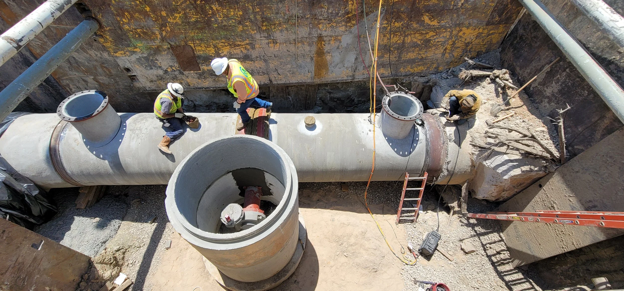 Construction workers installing large underground pipes within a deep excavation site, with tools and safety equipment visible.