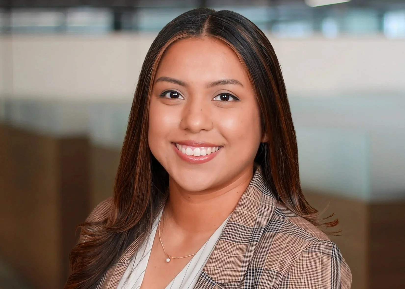 A professional woman with shoulder-length light brown hair, smiling, wearing a dark pinstripe suit and a gold necklace, in an office setting with glass partitions.