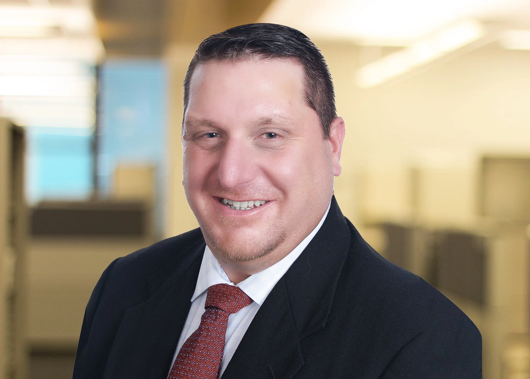 A smiling man in a gray suit, white shirt, and maroon tie, standing in a hallway with framed pictures on the wall.