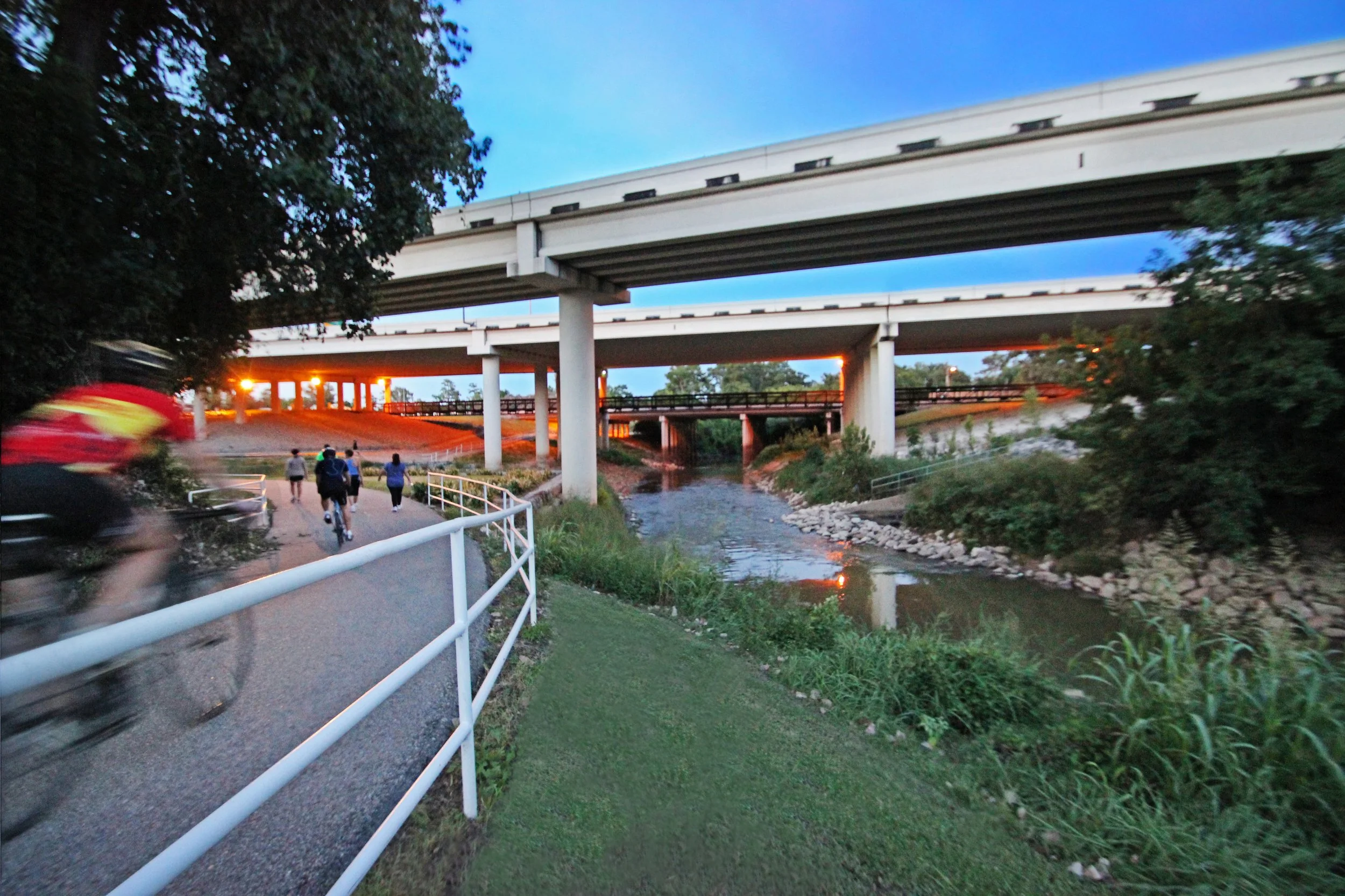 People walking, biking, and running on a trail under a large overpass bridge with a creek flowing underneath during dusk.