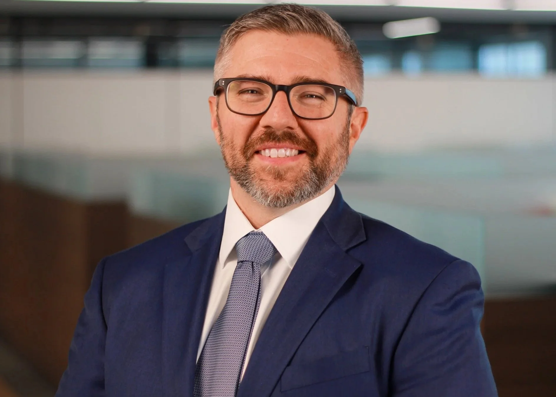 A middle-aged man with short gray hair, glasses, and a beard, wearing a navy blue suit, white shirt, and gray tie, smiling confidently in a modern office environment.