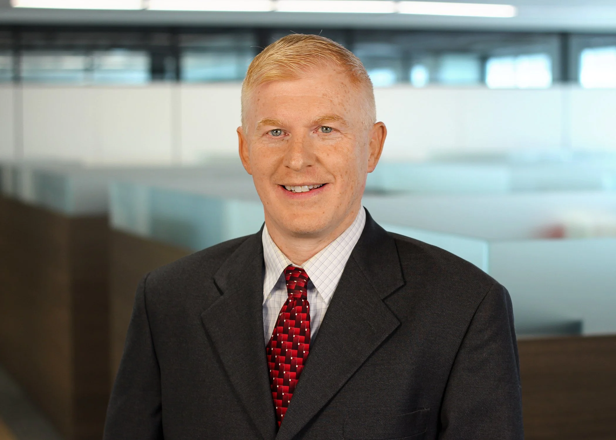 Portrait of a man in a business suit smiling in an office setting with glass walls.