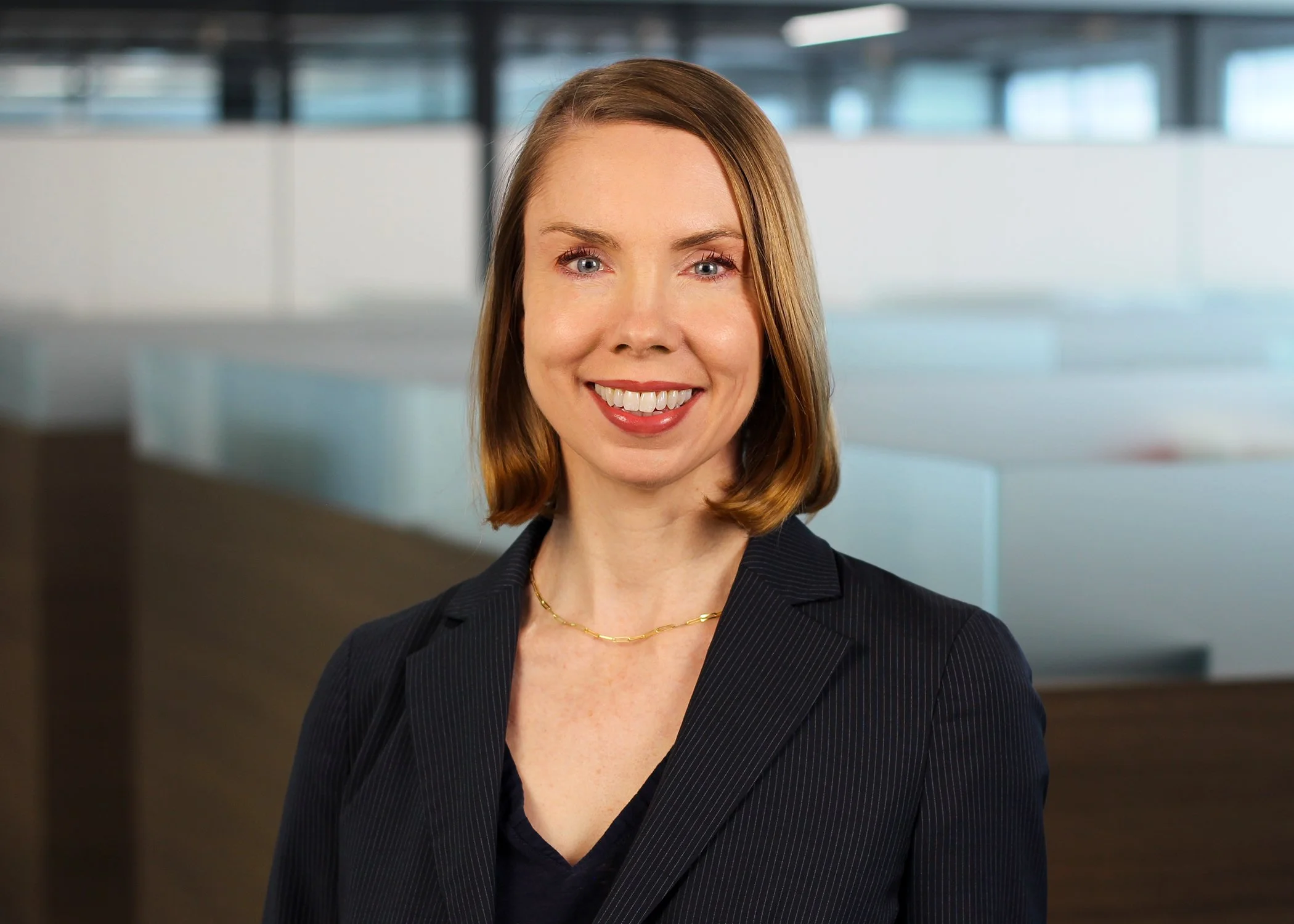 A professional woman with shoulder-length light brown hair, smiling, wearing a dark pinstripe suit and a gold necklace, in an office setting with glass partitions.