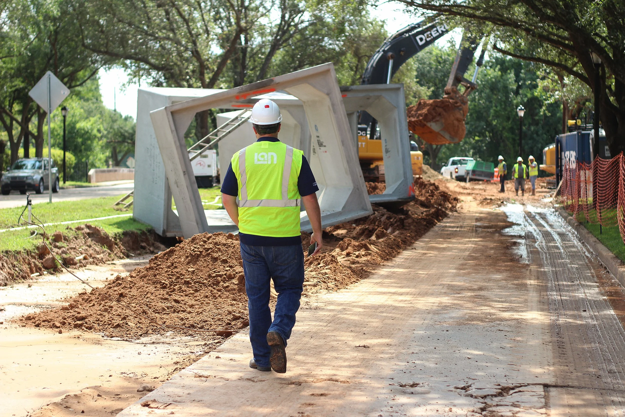 Construction workers wearing safety vests and helmets work on a sidewalk replacement project. One worker is walking on the sidewalk, while others are in the background near construction equipment and a large excavator digging up ground. The scene is set in a park-like area with trees and cars in the distance.