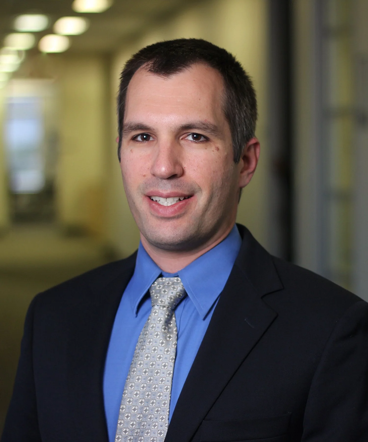 Professional headshot of a man in a business suit, wearing a blue shirt and a gray patterned tie, standing in an office corridor with yellow walls and windows.