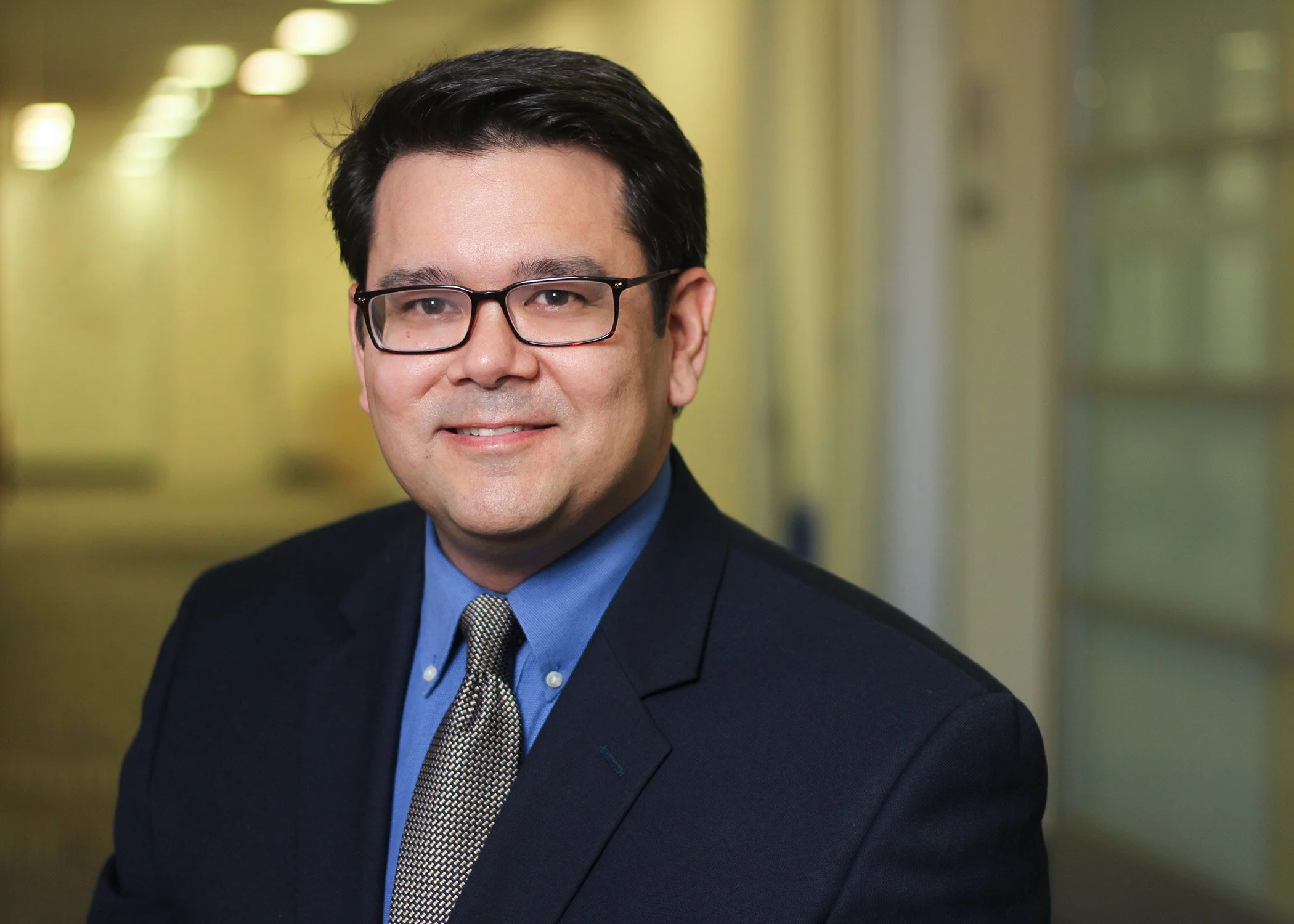 Professional headshot of a man in a business suit, wearing a blue shirt and a gray patterned tie, standing in an office corridor with yellow walls and windows.