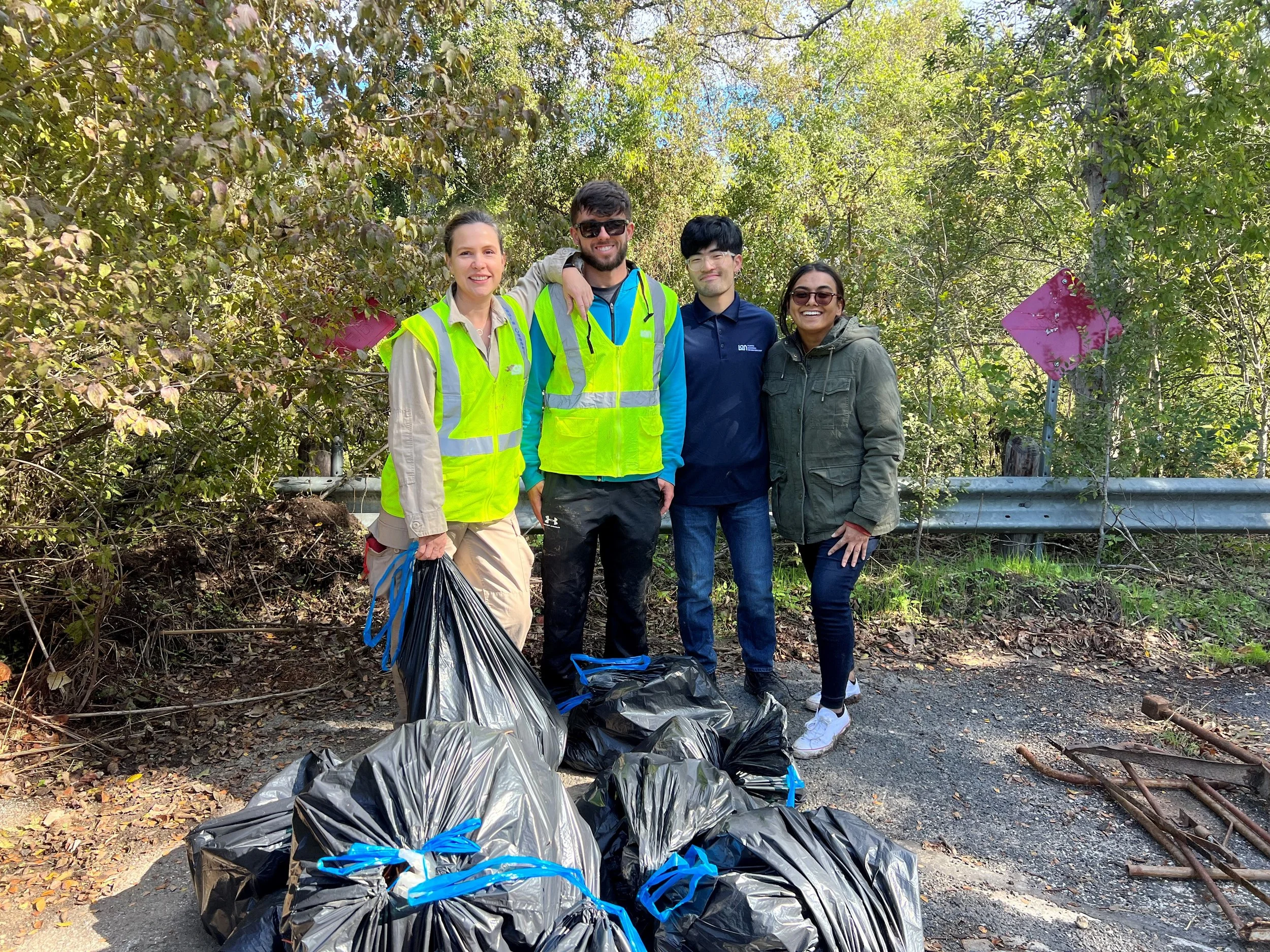 Four people standing outdoors in front of bushes, smiling, with large trash bags filled with collected litter on the ground in front. They are wearing casual clothes and some are in high-visibility vests. It appears to be a community cleanup event on a sunny day.