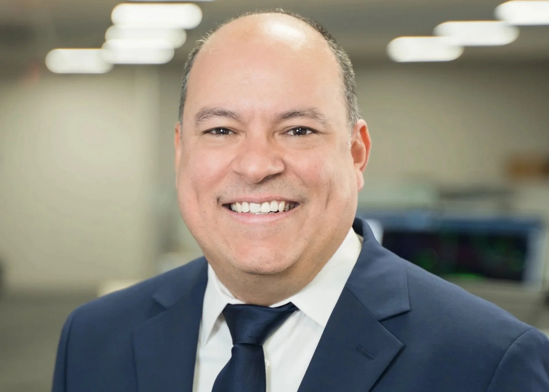 Professional headshot of a man in a business suit, wearing a blue shirt and a gray patterned tie, standing in an office corridor with yellow walls and windows.