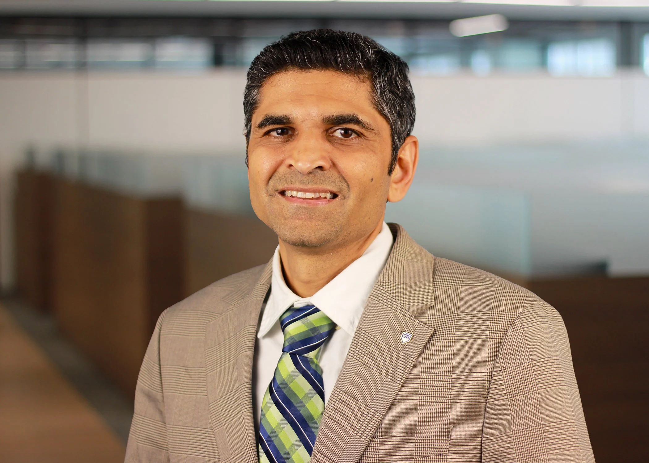 A smiling man in a gray suit, white shirt, and maroon tie, standing in a hallway with framed pictures on the wall.