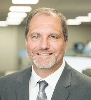 Portrait of a man in a business suit smiling in an office setting with glass walls.