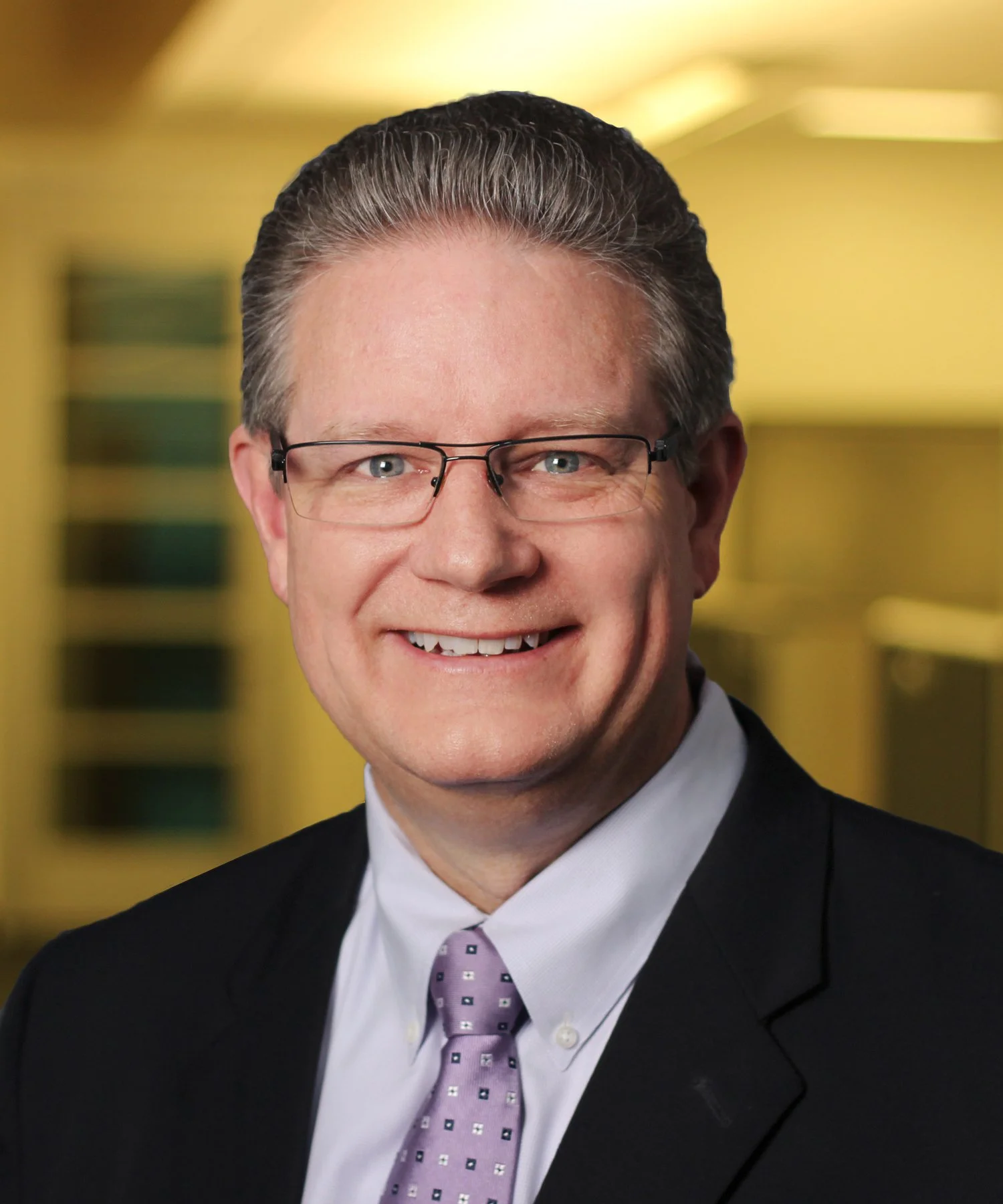 Professional headshot of a man in a business suit, wearing a blue shirt and a gray patterned tie, standing in an office corridor with yellow walls and windows.