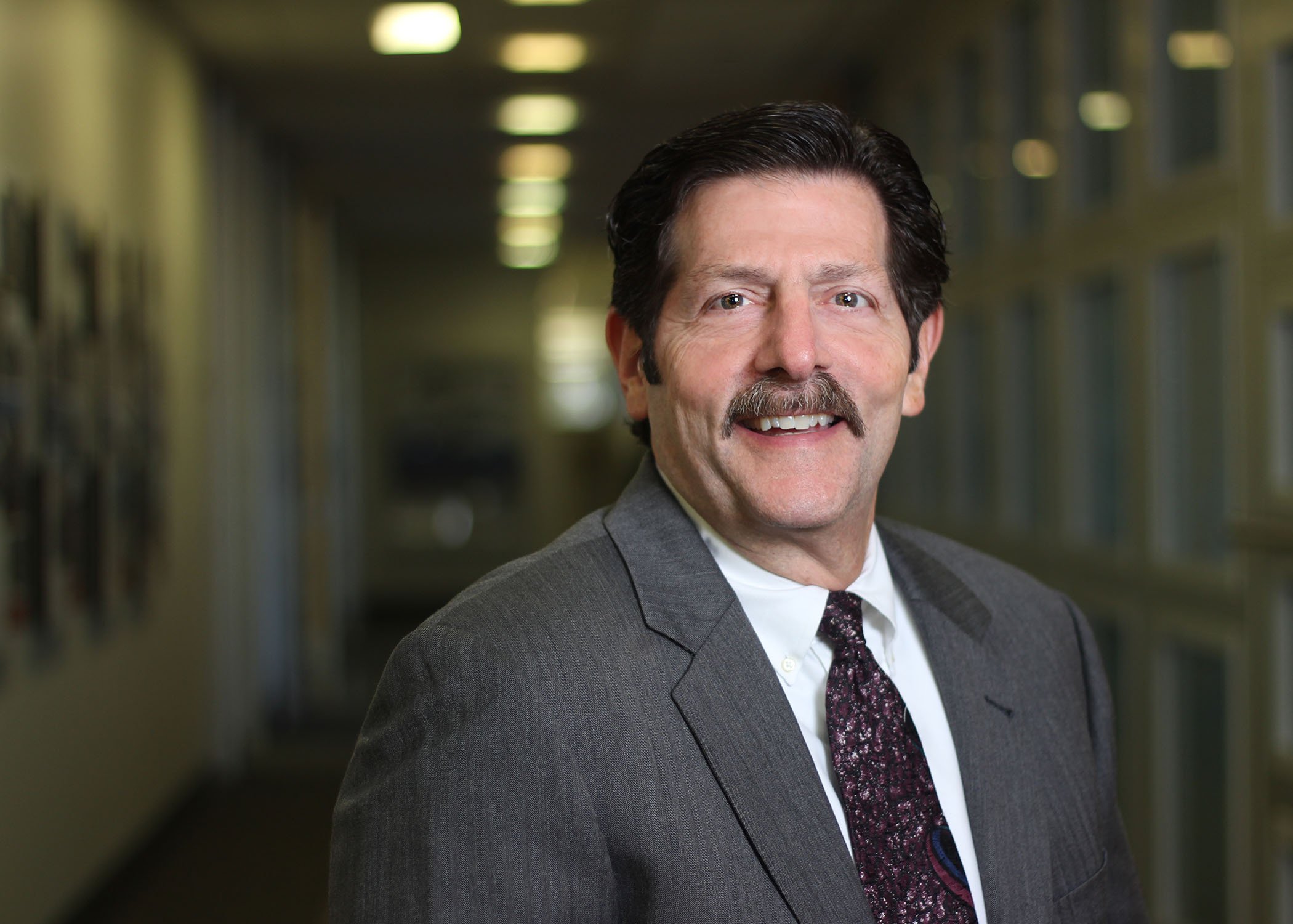 Man with brown hair and mustache, wearing a grey suit with a colorful tie, standing in a hallway with items hanging on the wall