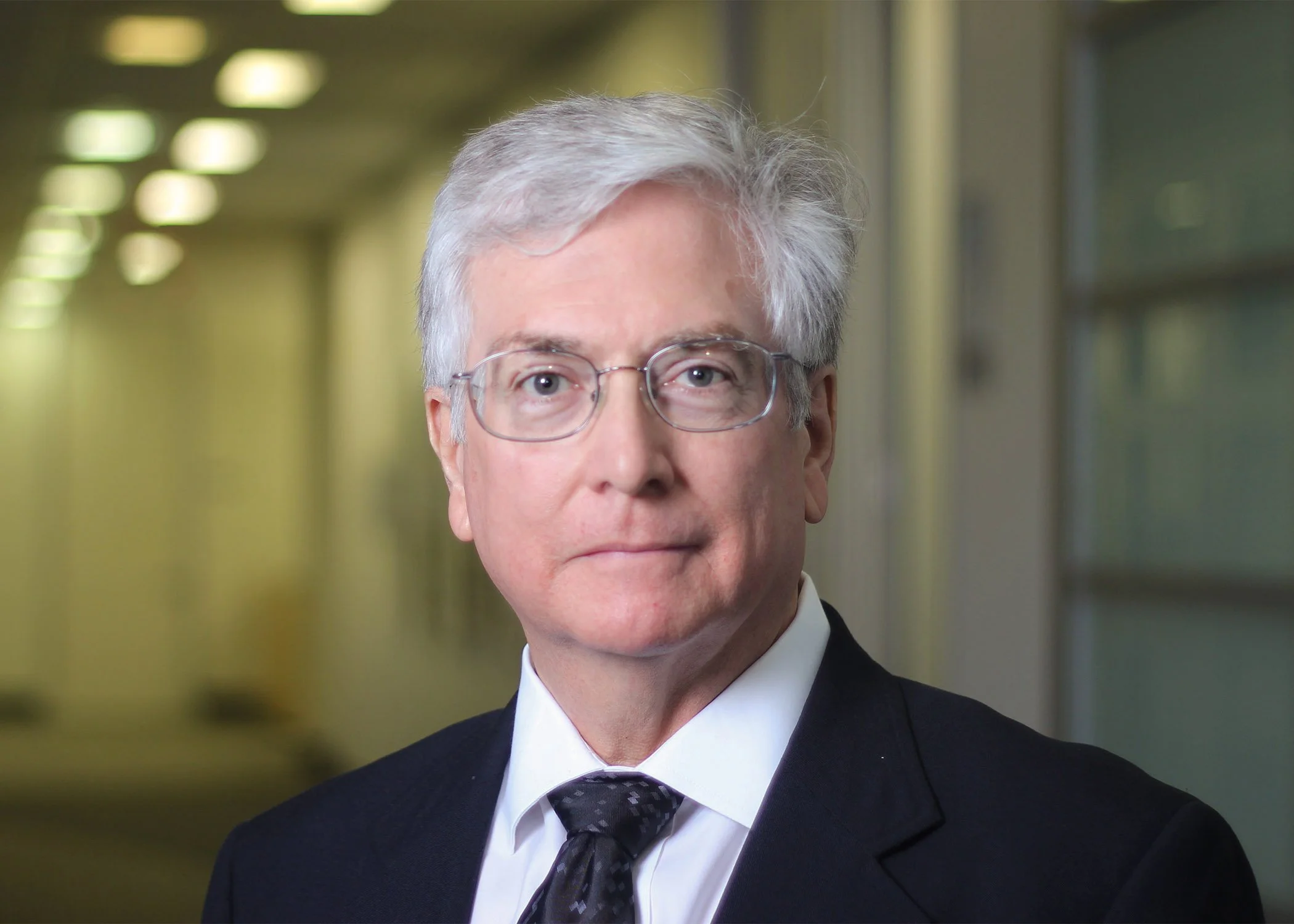 Portrait of a man with white hair and glasses in a business suit smiling in an office setting with glass walls.