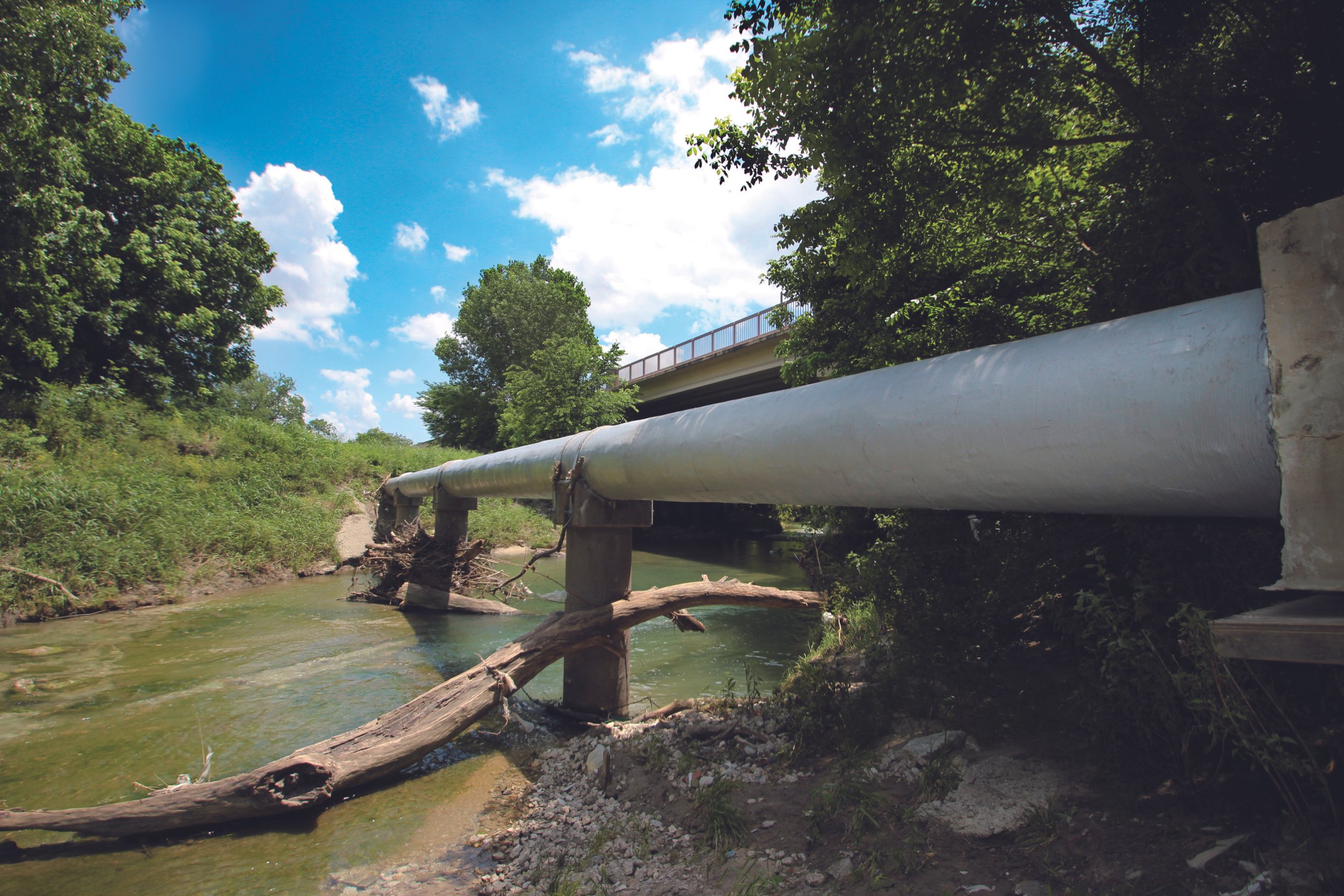 Large white pipeline running above a small creek, with trees and a bridge in the background, under a partly cloudy sky.