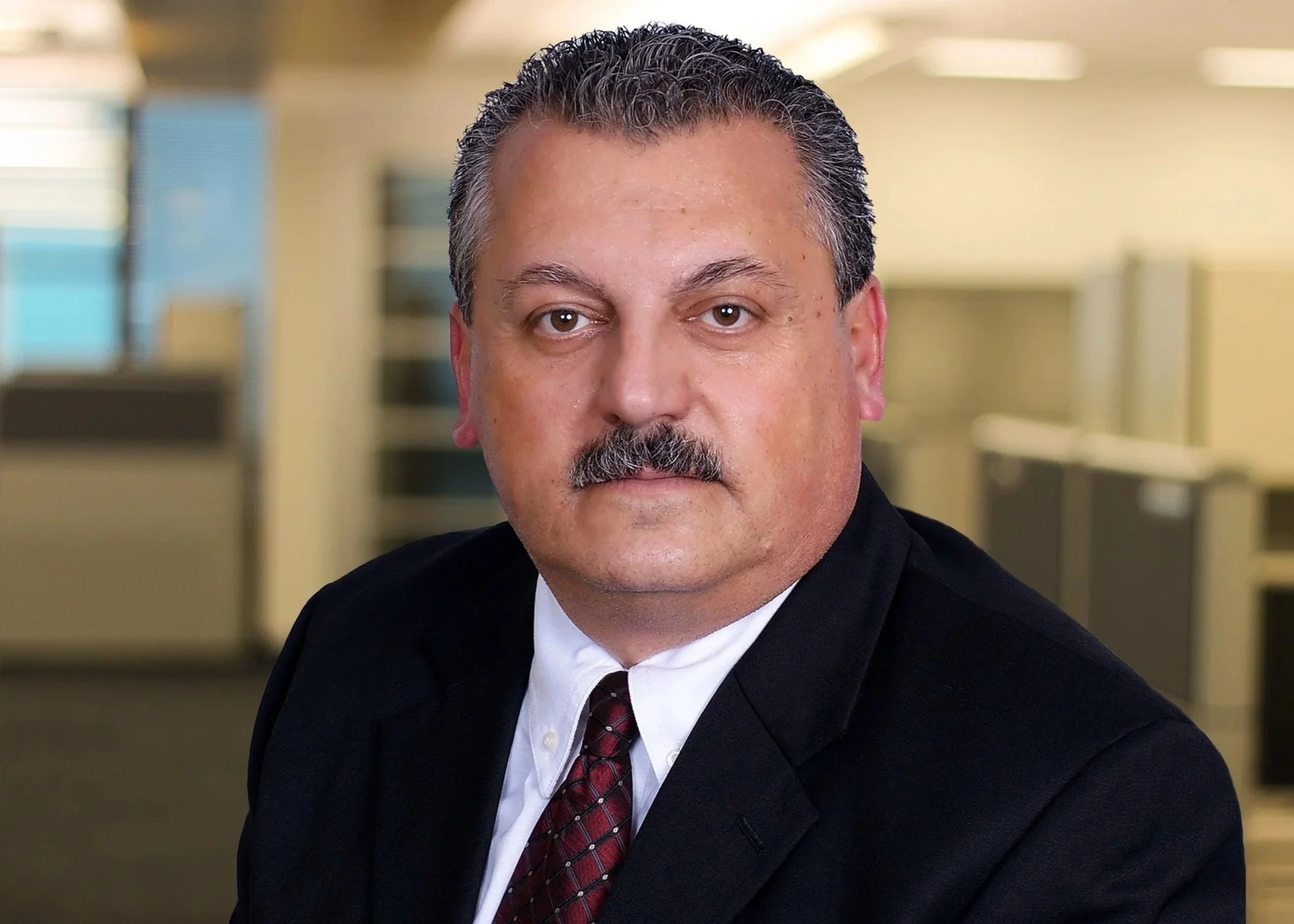 A middle-aged man with dark hair, a mustache, wearing a dark suit, white shirt, and maroon tie posed in an office setting.