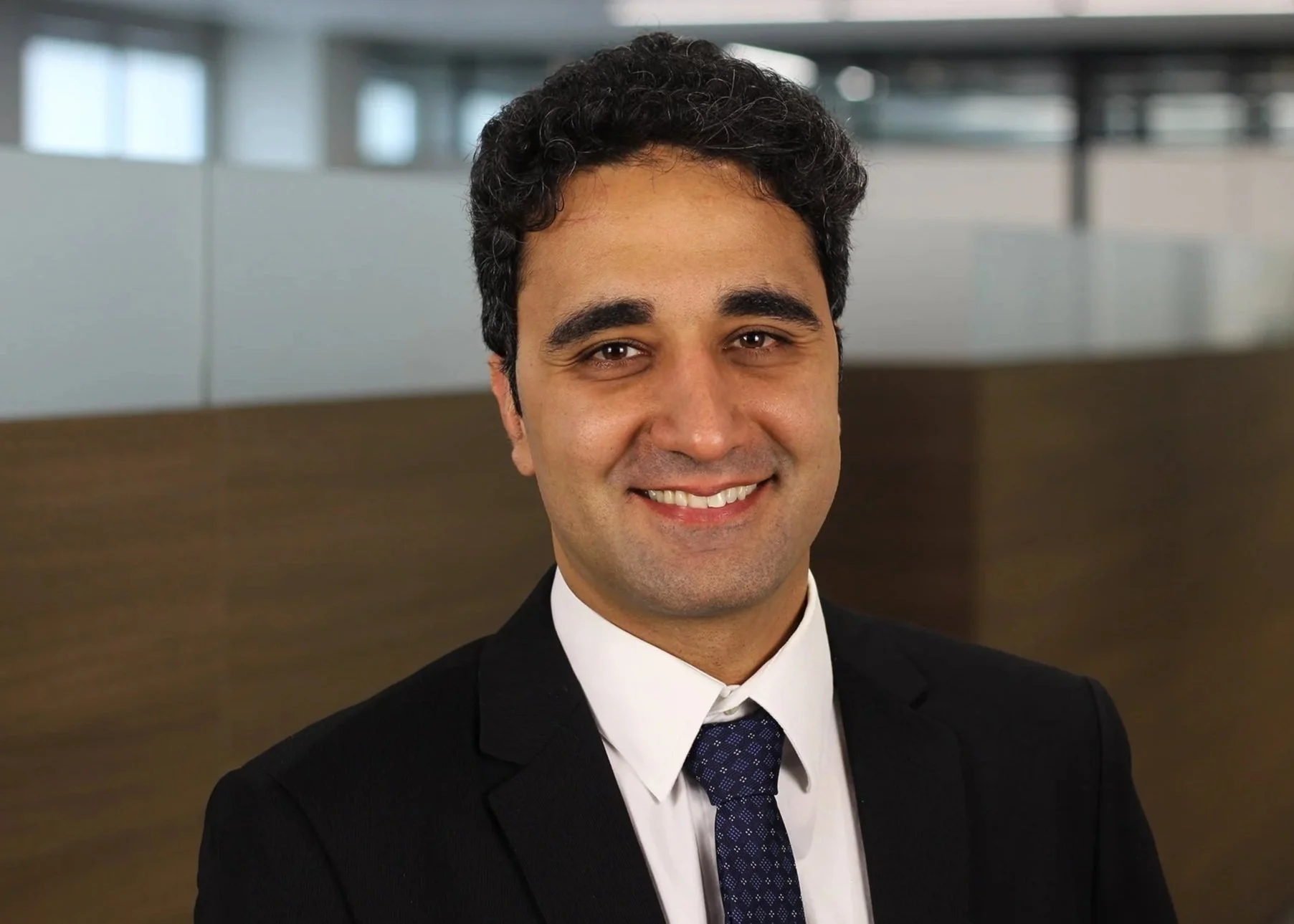 Portrait of a man in a business suit smiling in an office setting with glass walls.
