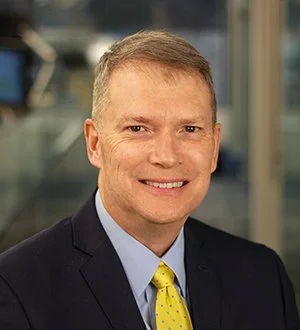 A smiling man in a gray suit, white shirt, and maroon tie, standing in a hallway with framed pictures on the wall.