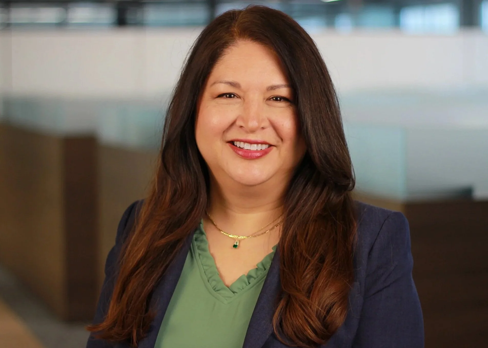 A woman with long brown hair, wearing a green top and a navy blazer, smiling in an indoor office setting.