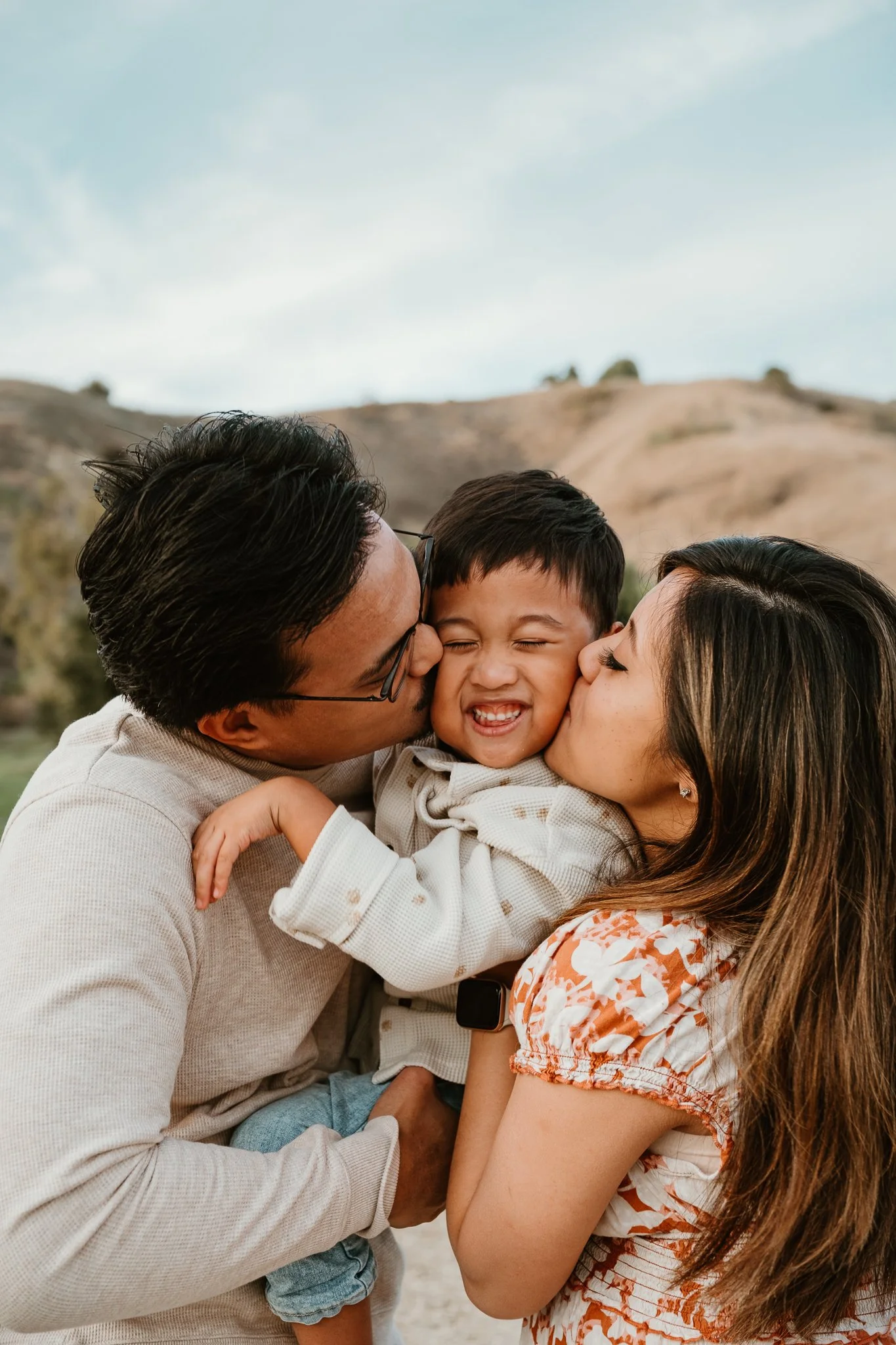 A family of three shows affection outdoors, with a man and woman kissing their smiling young son on each cheek, in front of a hilly landscape.