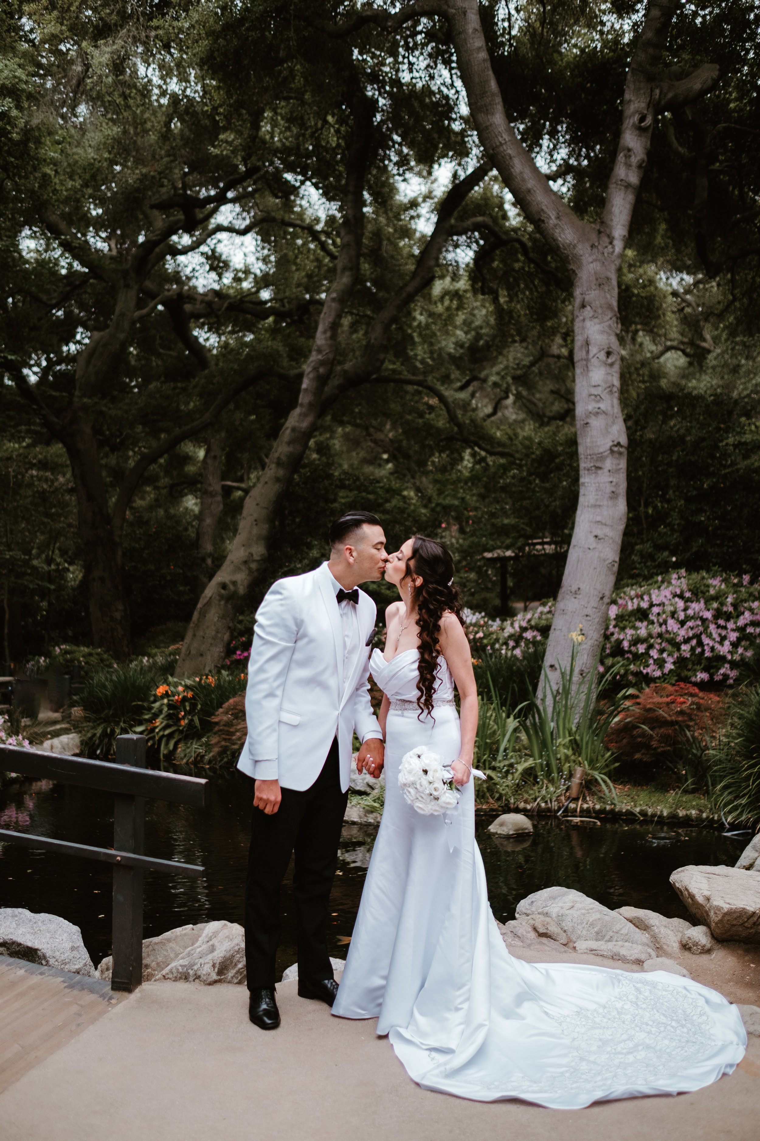 A bride and groom kissing in a garden with trees, flowers, and a pond, during their wedding.