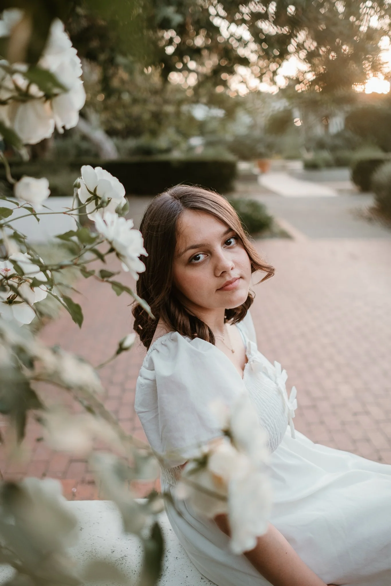 A young woman with brown hair and a white dress sitting outdoors on a bench, surrounded by white flowers with a sunset in the background.