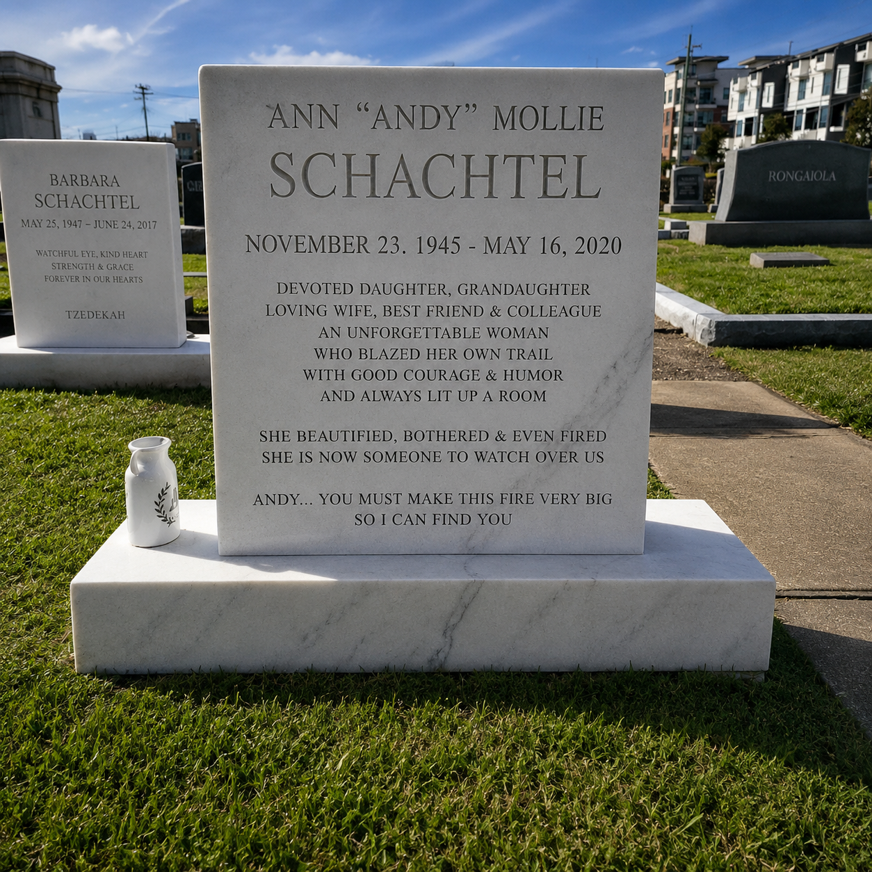 Gravestone for Ann "Andy" Mollie Schachtel with a small white vase beside it, situated in a cemetery with other headstones and apartment buildings in the background, clear sky above.