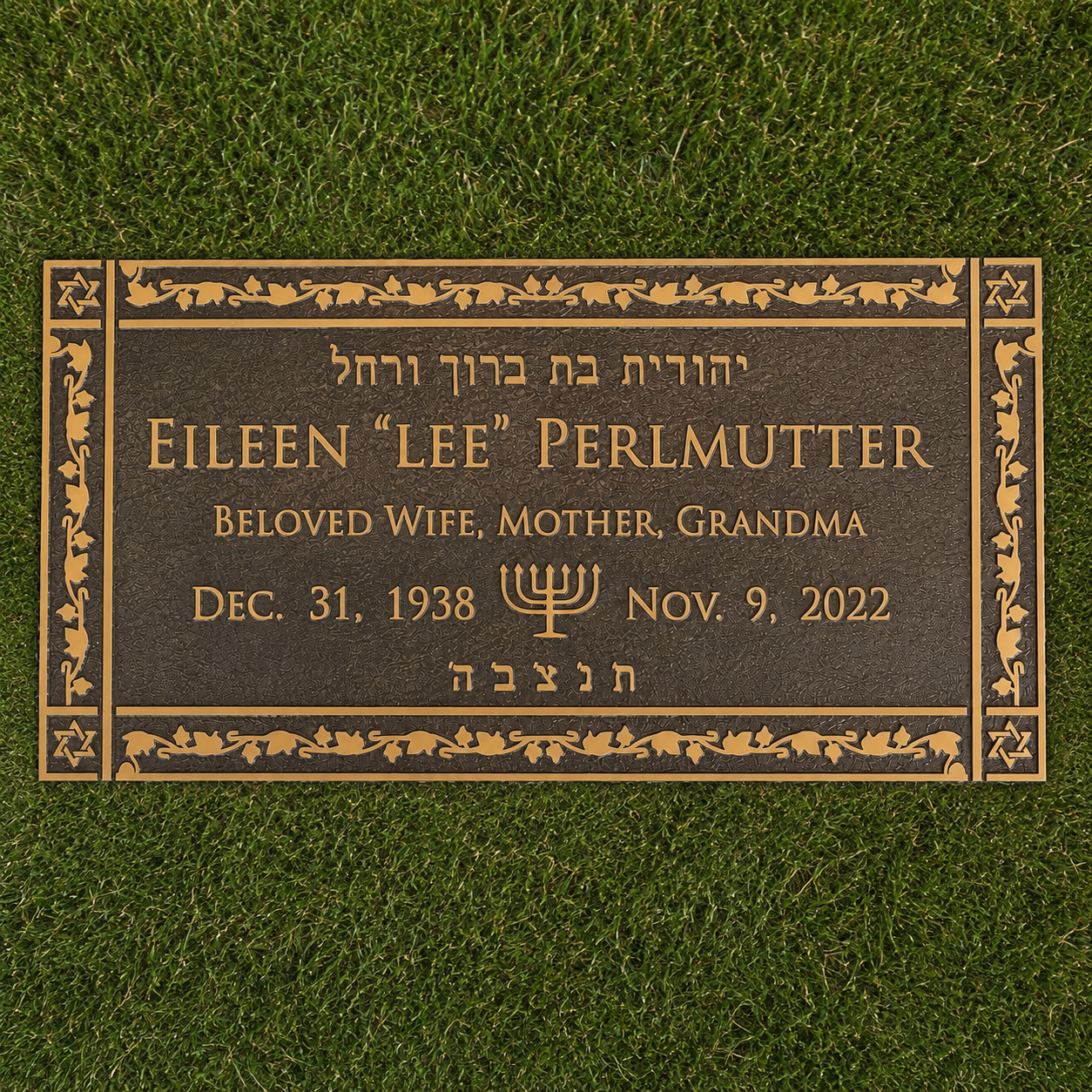 A bronze grave marker on green grass dedicated to Eileen 'Lee' Perlmutter, who was a beloved wife, mother, and grandma, born December 31, 1938, and passed away on November 9, 2022. The memorial features a menorah symbol and Hebrew inscription.