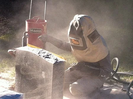 Person wearing protective gear using a power tool to cut a piece of stone or concrete outdoors.