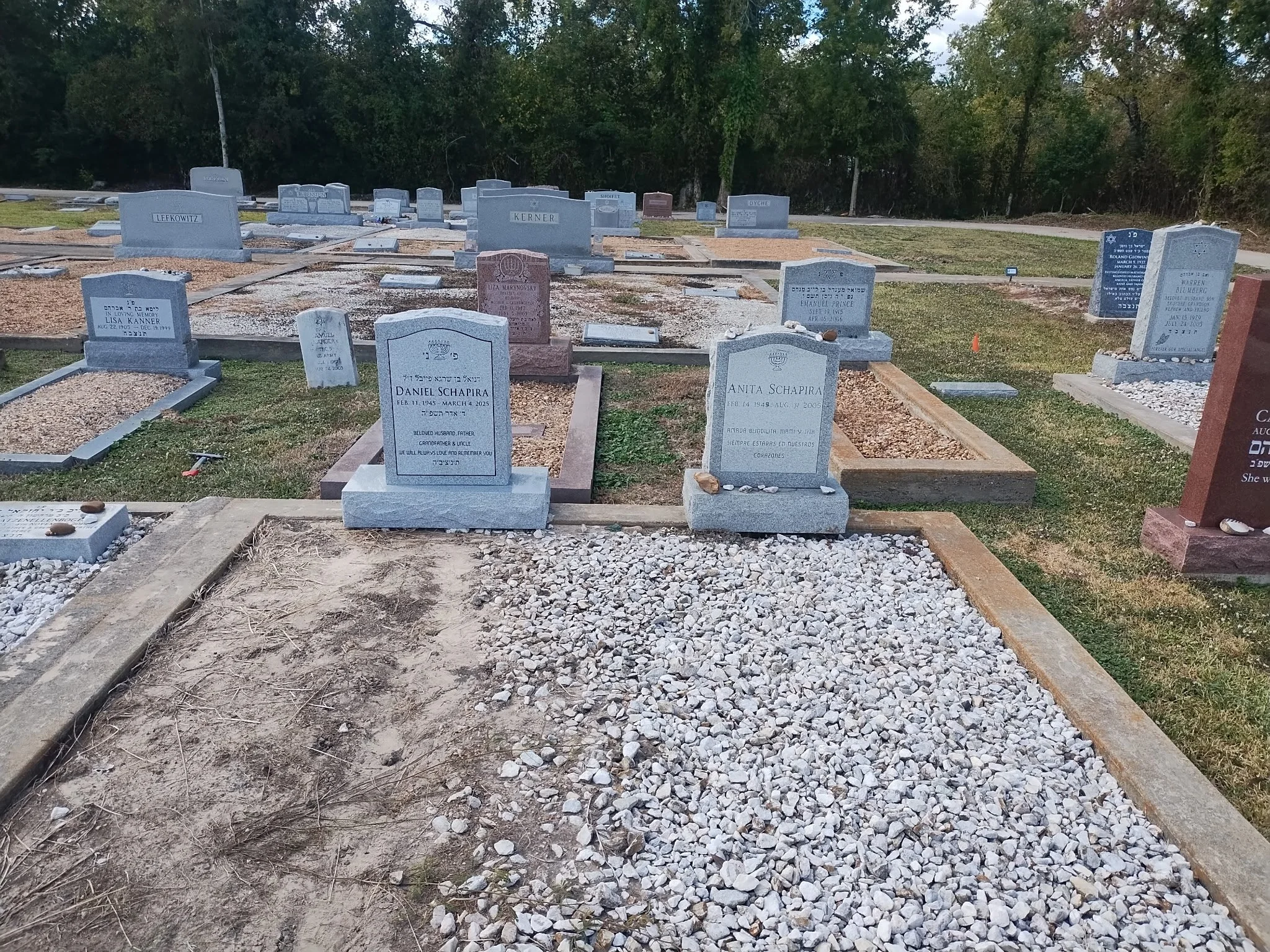 A cemetery with numerous headstones, some with inscriptions in English and Hebrew, surrounded by trees and a curved path, with a gravel pathway in the foreground.