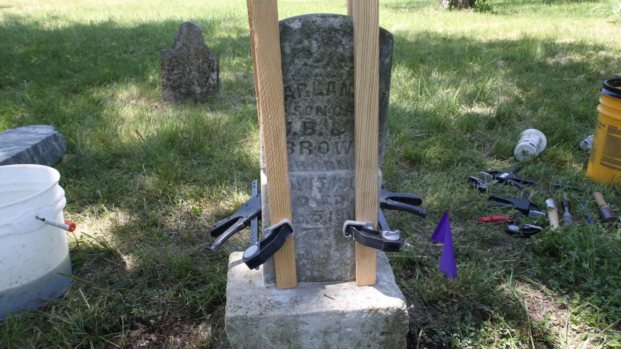 Restoration of a stone grave marker with wooden supports and clamps in a grassy outdoor cemetery, surrounded by tools and containers.