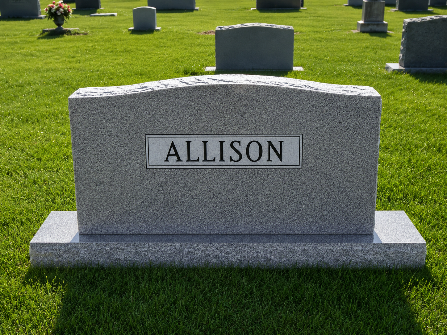 Gray granite tombstone with the name Allison engraved, located in a green cemetery with other headstones and a flower arrangement in a vase in the background.
