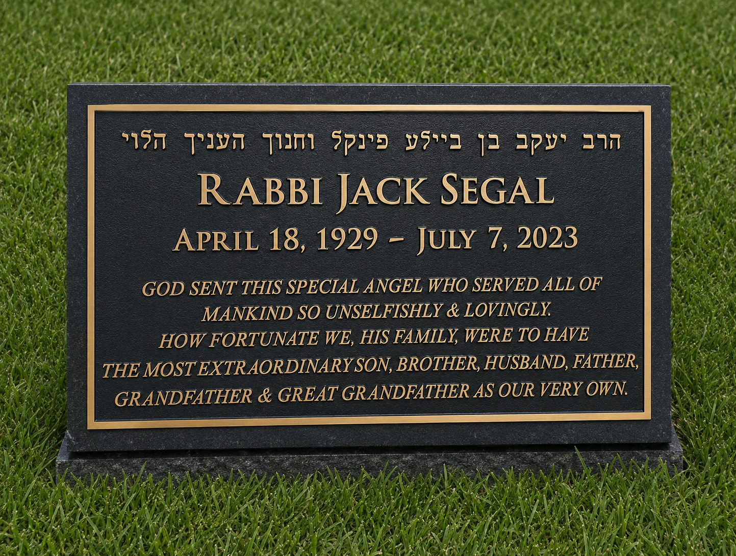 A black memorial plaque with gold borders and lettering on green grass. It honors Rabbi Jack Segal, who was born April 18, 1929, and died July 7, 2023. The plaque includes a message about his service as a special angel sent by God, who served mankind selflessly and lovingly, and recognized as an extraordinary son, brother, husband, father, grandfather, and great-grandfather.