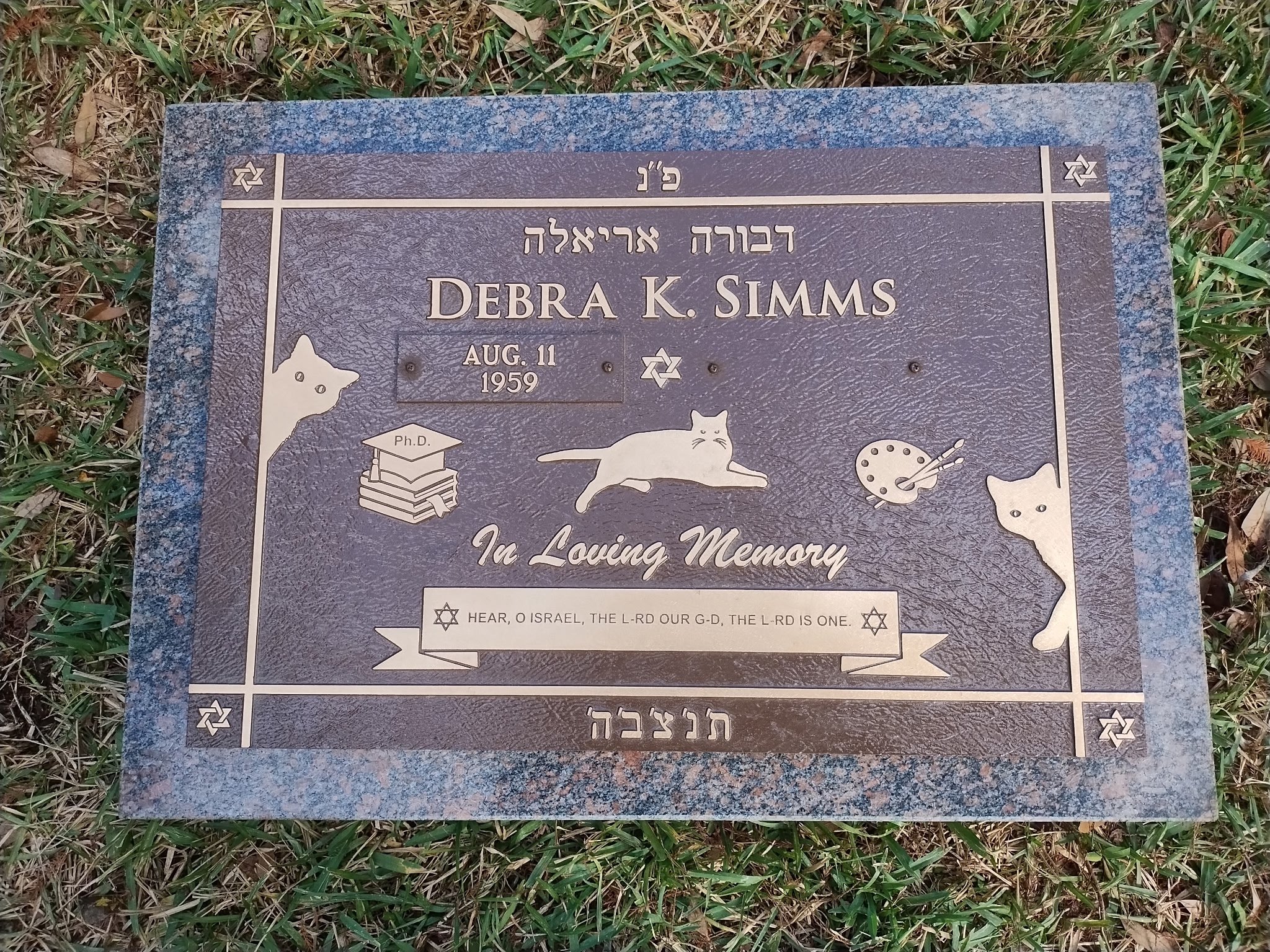 Gravestone plaque for Debra K. Simms, with decorative elements including dog illustrations, books with a graduate cap, and a paint palette, situated on grass.