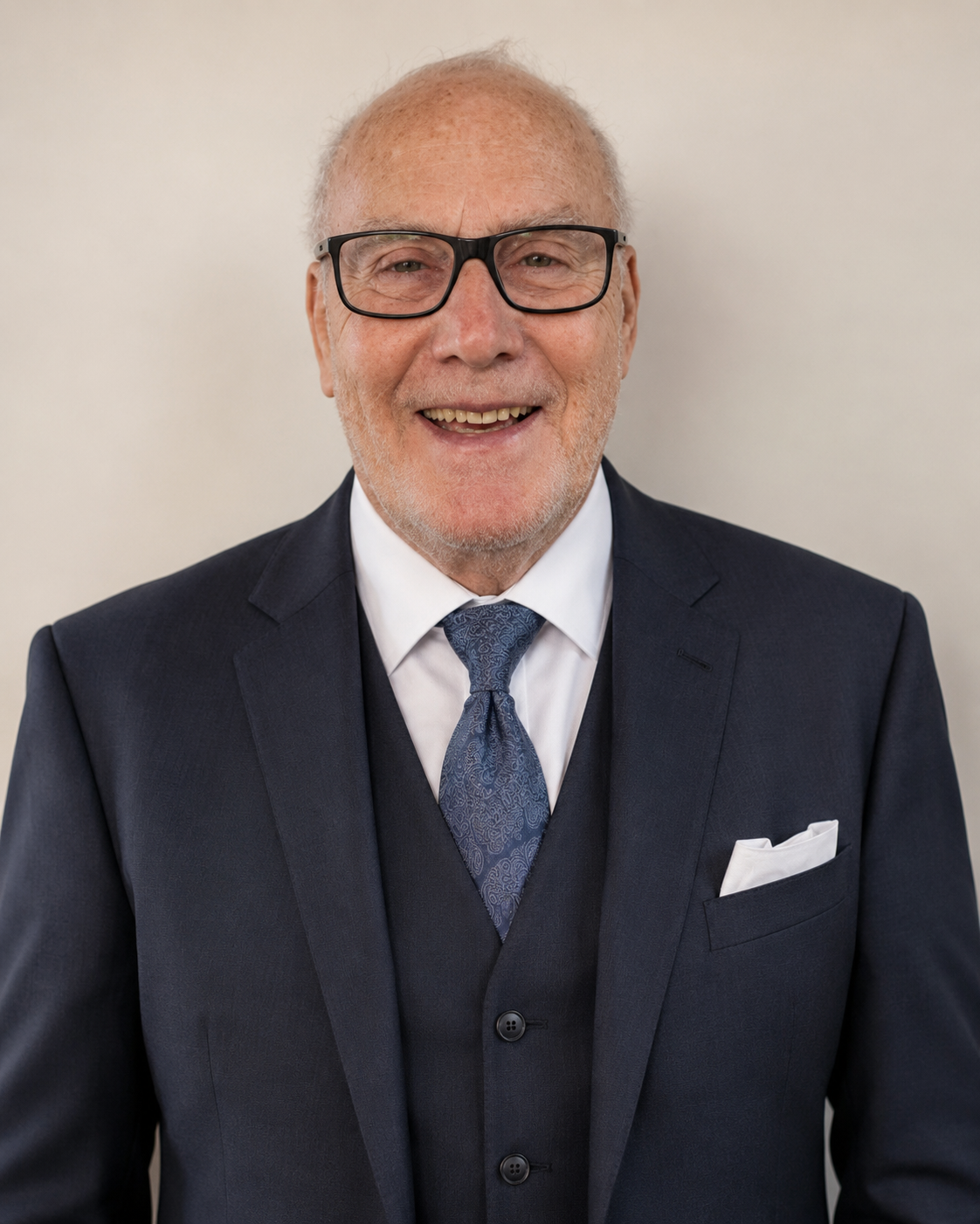 A smiling older man with glasses wearing a dark suit, white shirt, and a patterned blue tie, against a plain light background.